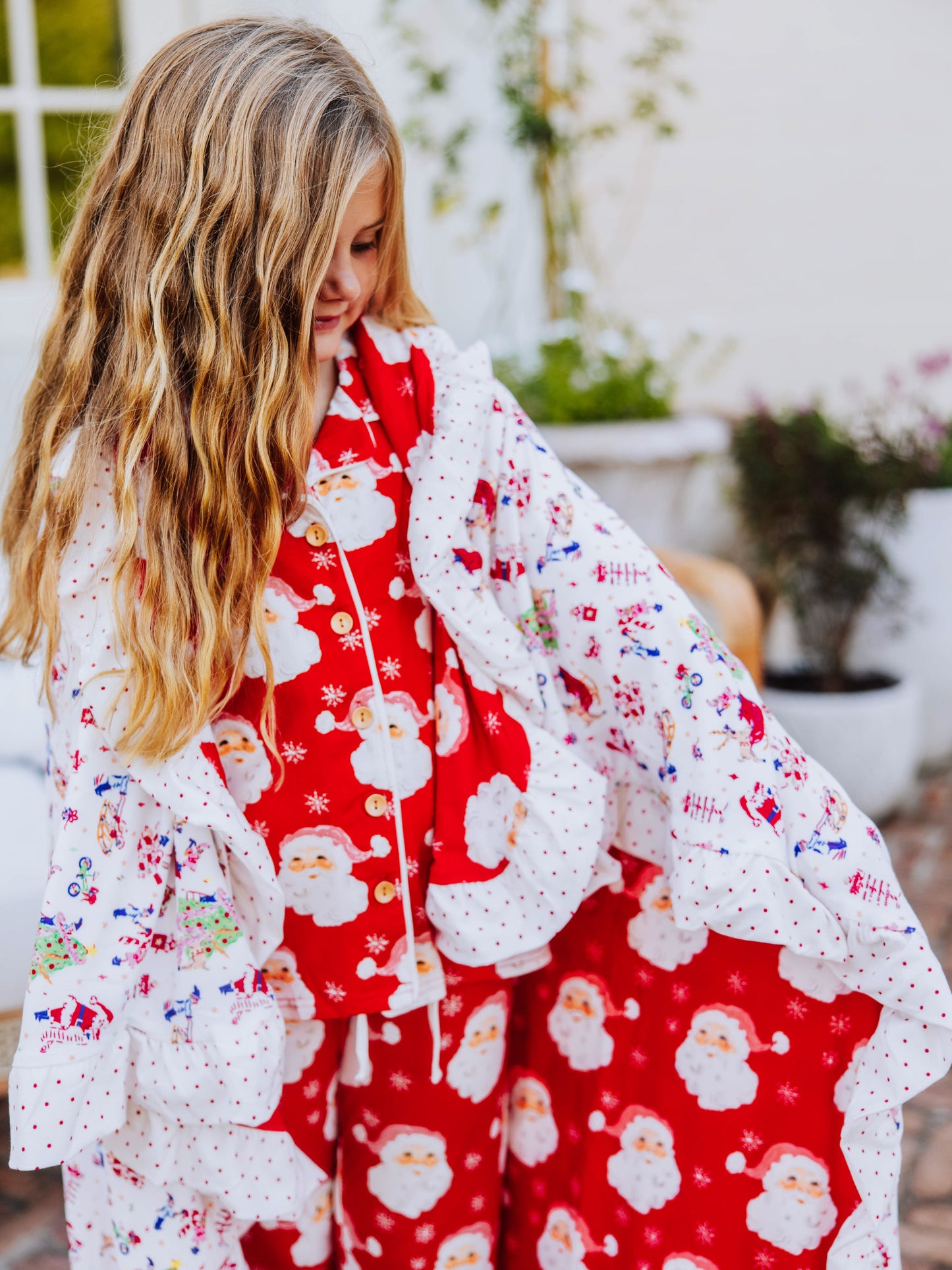 Girl wearing santa pajamas has a blanket wrapped around her. blanket has red santas and north pole prints on it.