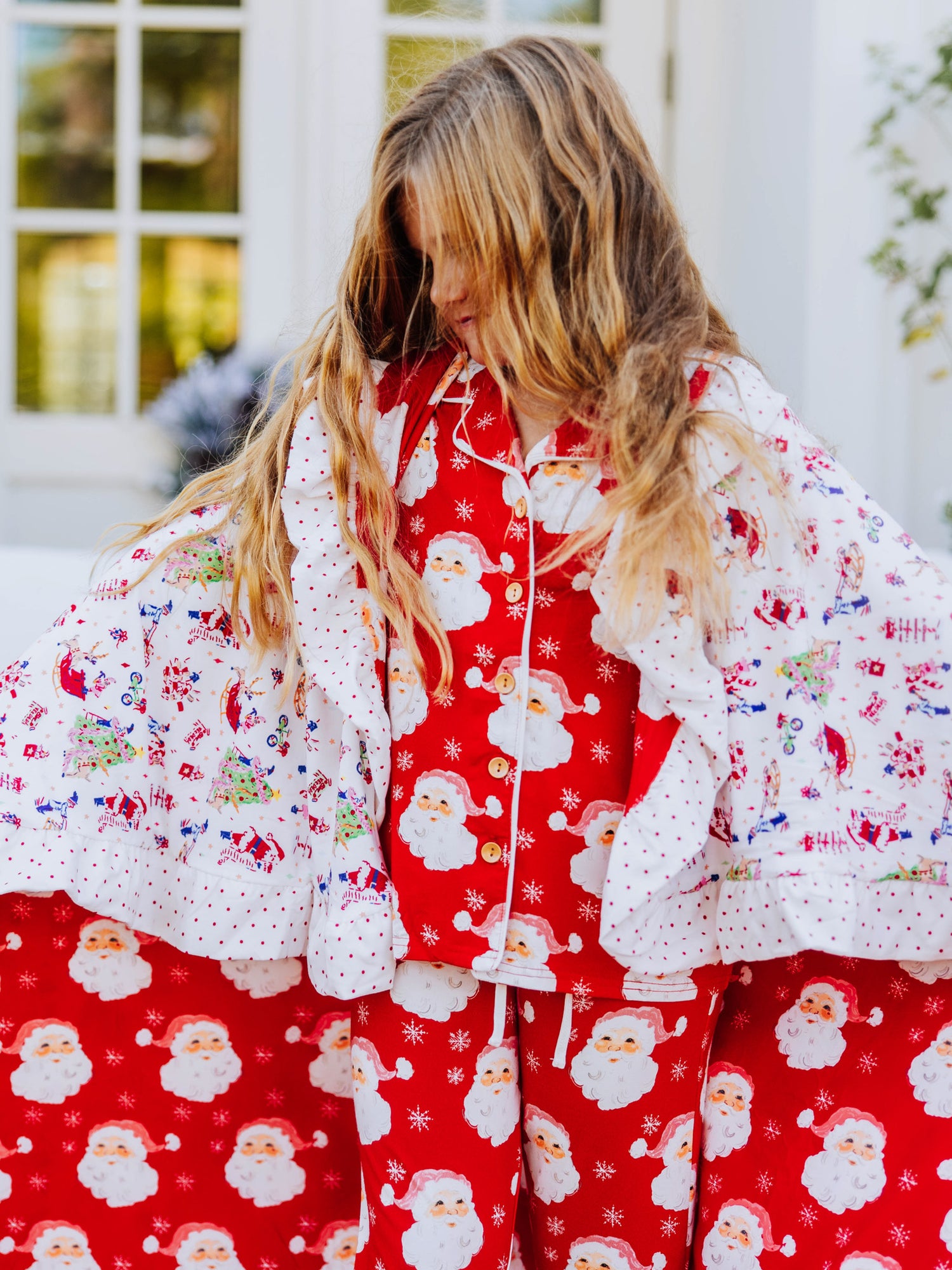 Girl wearing santa pajamas has a blanket wrapped around her. blanket has red santas and north pole prints on it.