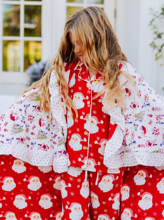 Girl wearing santa pajamas has a blanket wrapped around her. blanket has red santas and north pole prints on it.