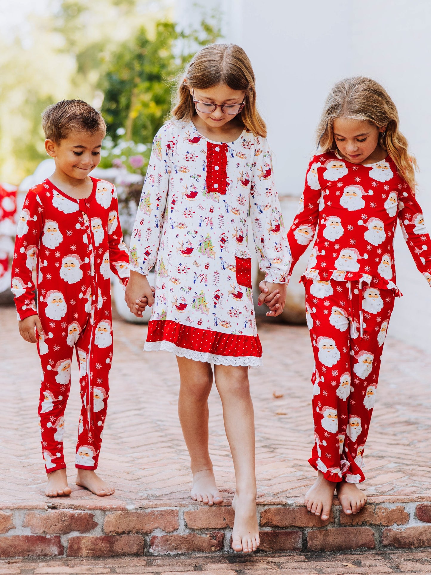Three models all holding hands and walking down the stairs. left model is a boy wearing a red santa layette, girl in the middle is wearing north pole printed play dress, and girl on the right is wearing matching print as boy.