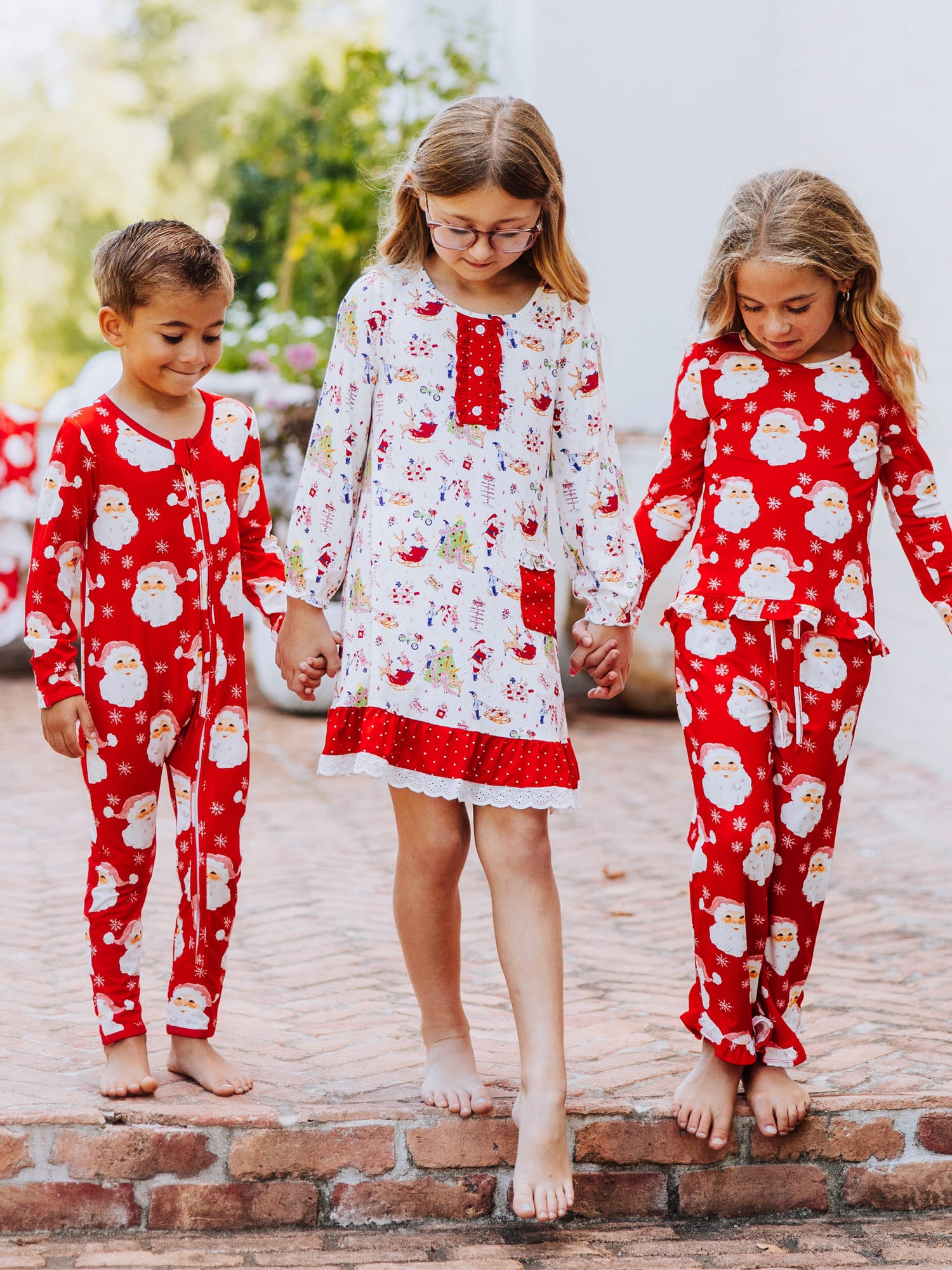 Three models all holding hands and walking down the stairs. left model is a boy wearing a red santa layette, girl in the middle is wearing north pole printed play dress, and girl on the right is wearing matching print as boy.