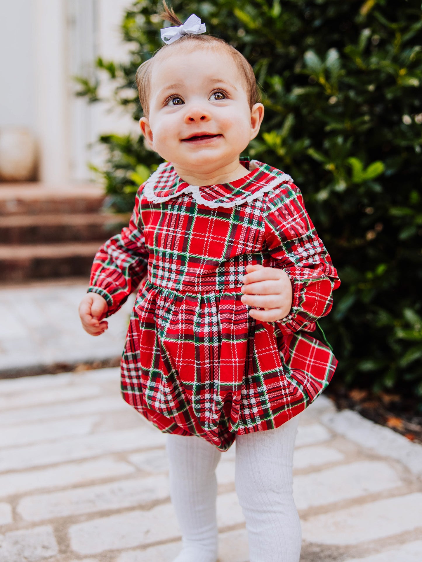 Toddler outside walking on steps in a traditional christmas plaid clothing bubble. Toddler is wearing ivory colored tights. 