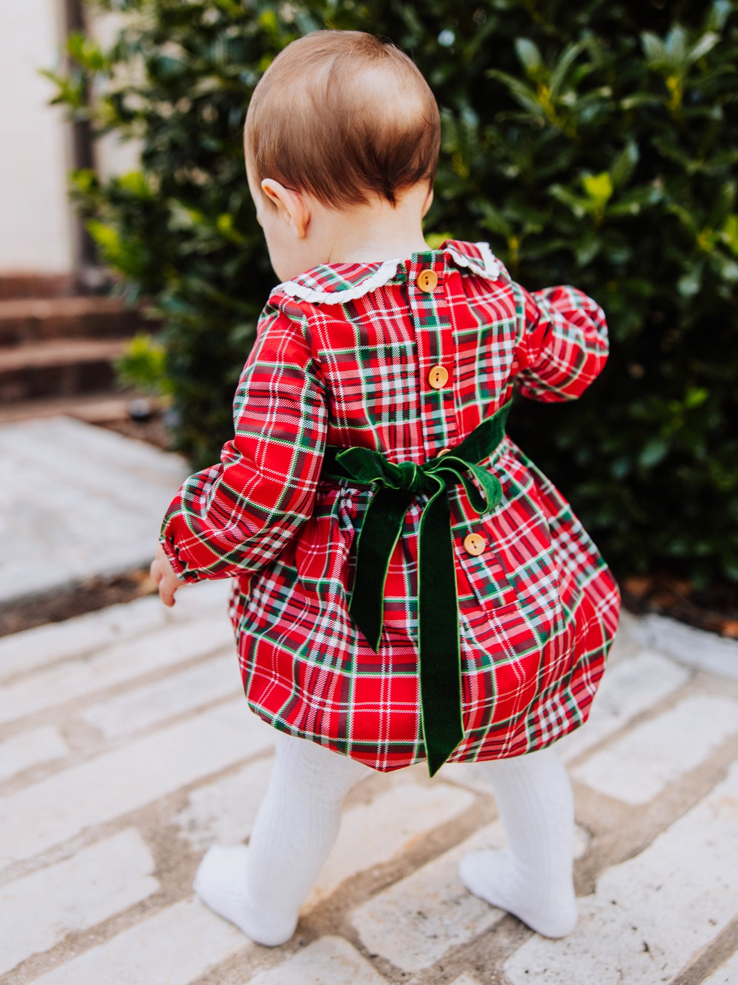 Close up of deep green velvet tie at back of traditional plaid colored bubble with wooden buttons. Toddler wearing ivory colored tights. 