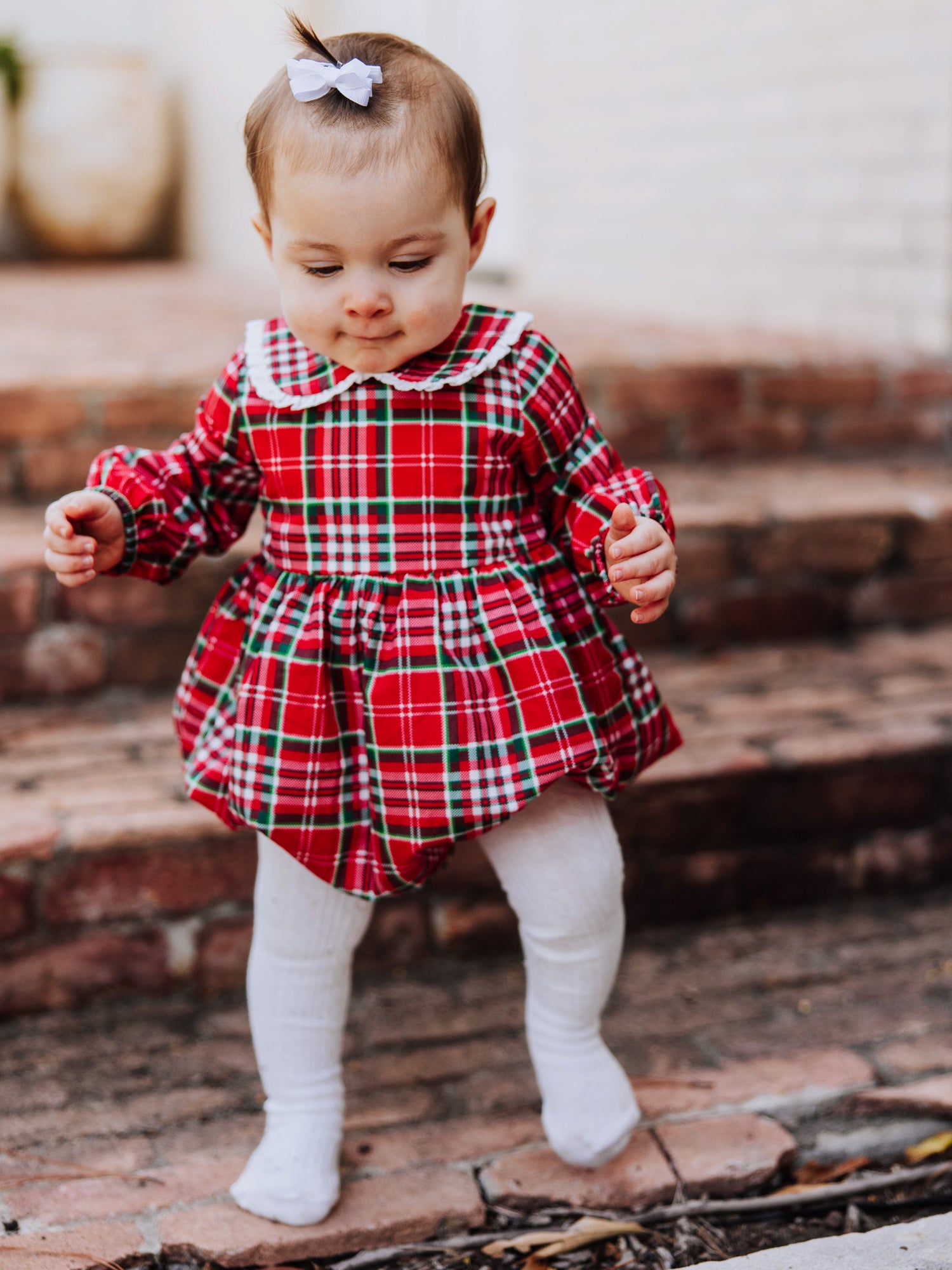oddler outside walking on steps in a traditional christmas plaid clothing bubble. Toddler is wearing ivory colored tights. 