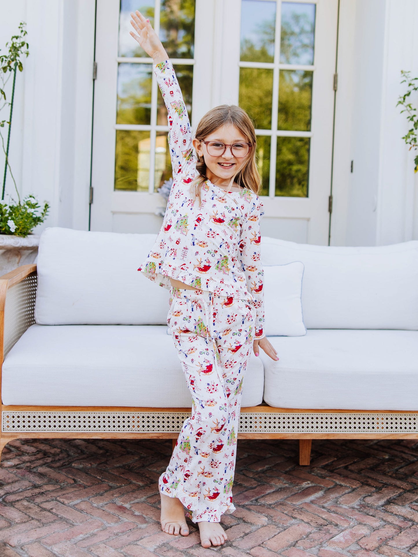 Girl standing in front of white couch posing with one hand up in the air. Girl is wearing a two piece matching ruffled pant set with a north pole print on it. 