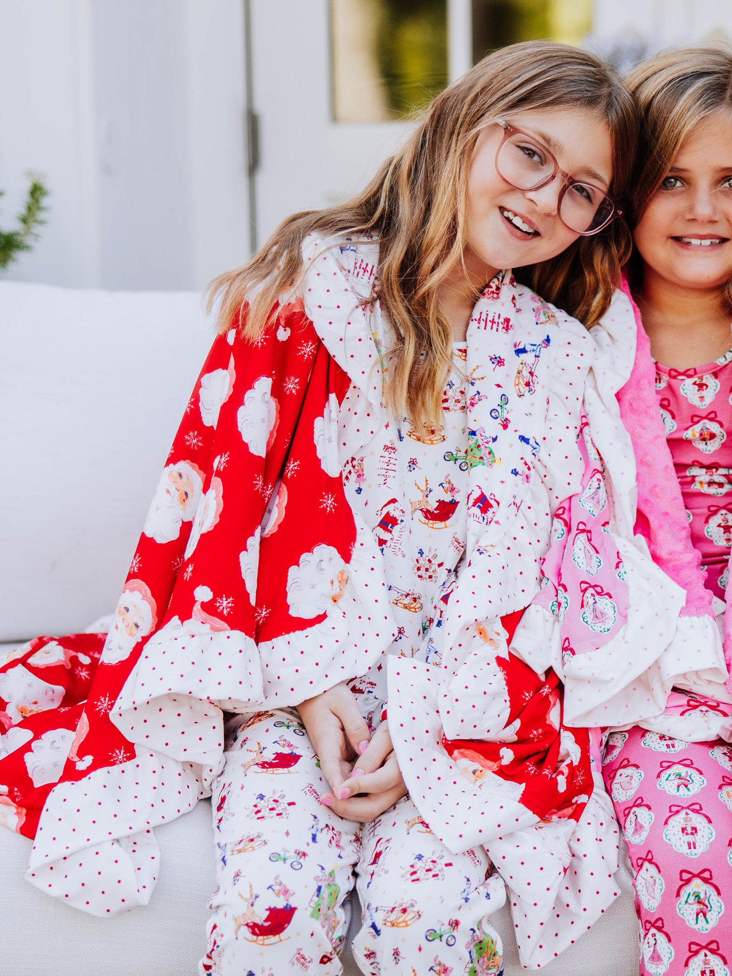 Girl sitting on couch with head against another girl. Both are wrapped up in Christmas blankets. 