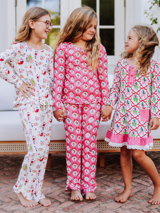 Three girls standing outside and holding hands. All are in different sets. Girl on left has hand on hip and is wearing a ruffled set that has north pole designs on it. Girl in middle is wearing a pink nutcracker set, and girl on the right is in a play dress with a multi-colored bow print all over. 