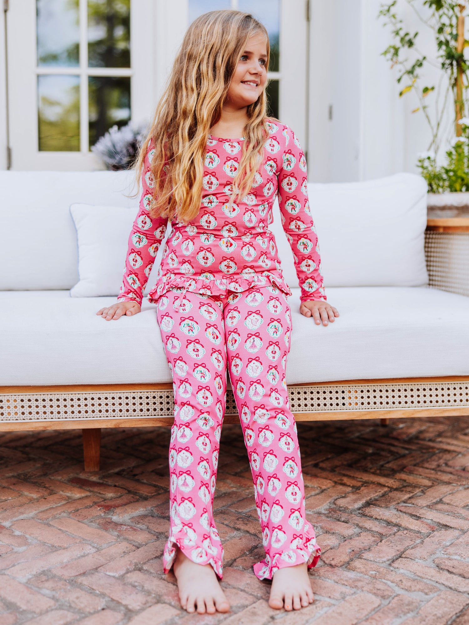 Girl sitting on edge of white couch wearing a two piece pink nutcracker ruffled pant set. 