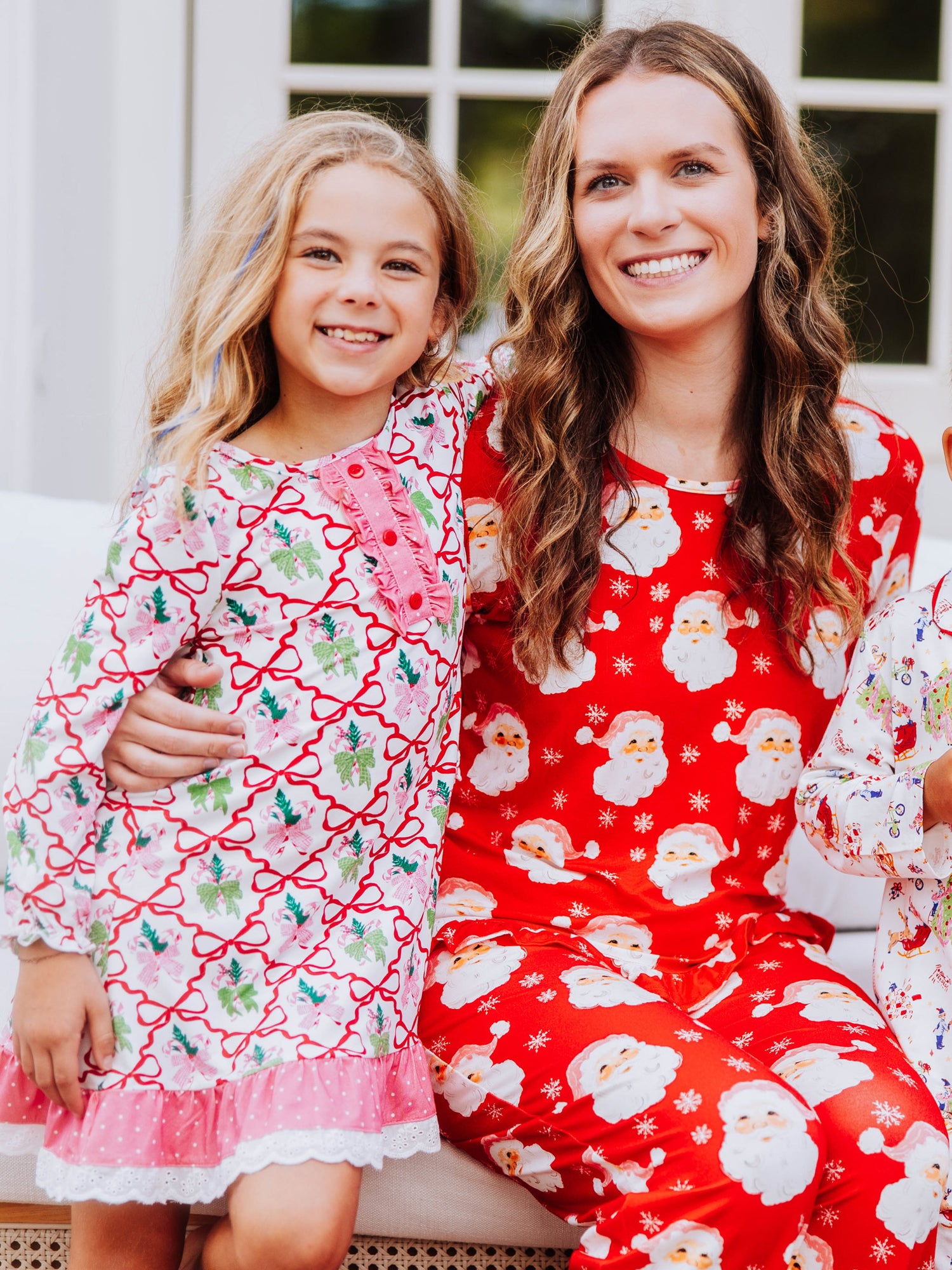 Girl and women sitting together and the women has her hand around girl. Girl is wearing a colorful bow printed dress and the women is wearing a red santa pair of pajamas. 