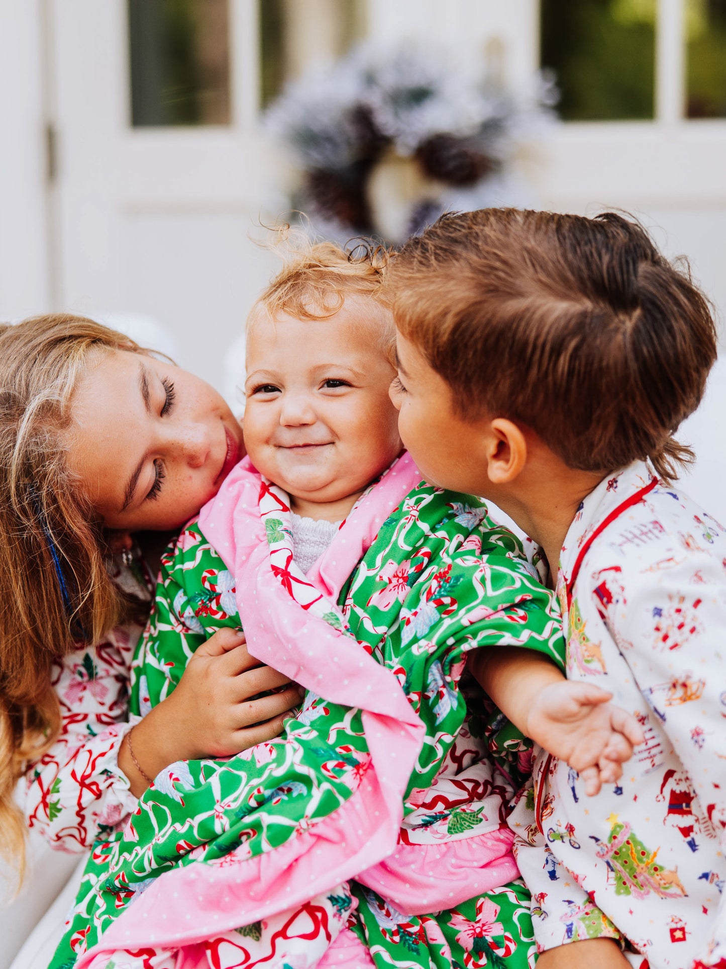 baby in the middle and girl on left and boy on right looking at baby. All have on christmas clothing and baby is wrapped in a blanket. 