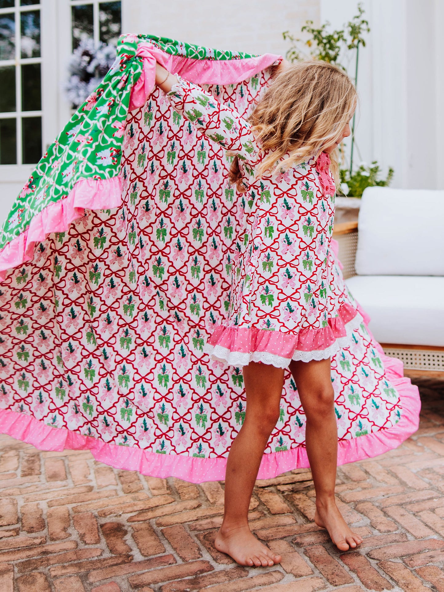 Girl holding out a colorful Christmas blanket over shoulder and is wearing a play dress that matches print on blanket. Print is colorful and has red bows throughout pattern.