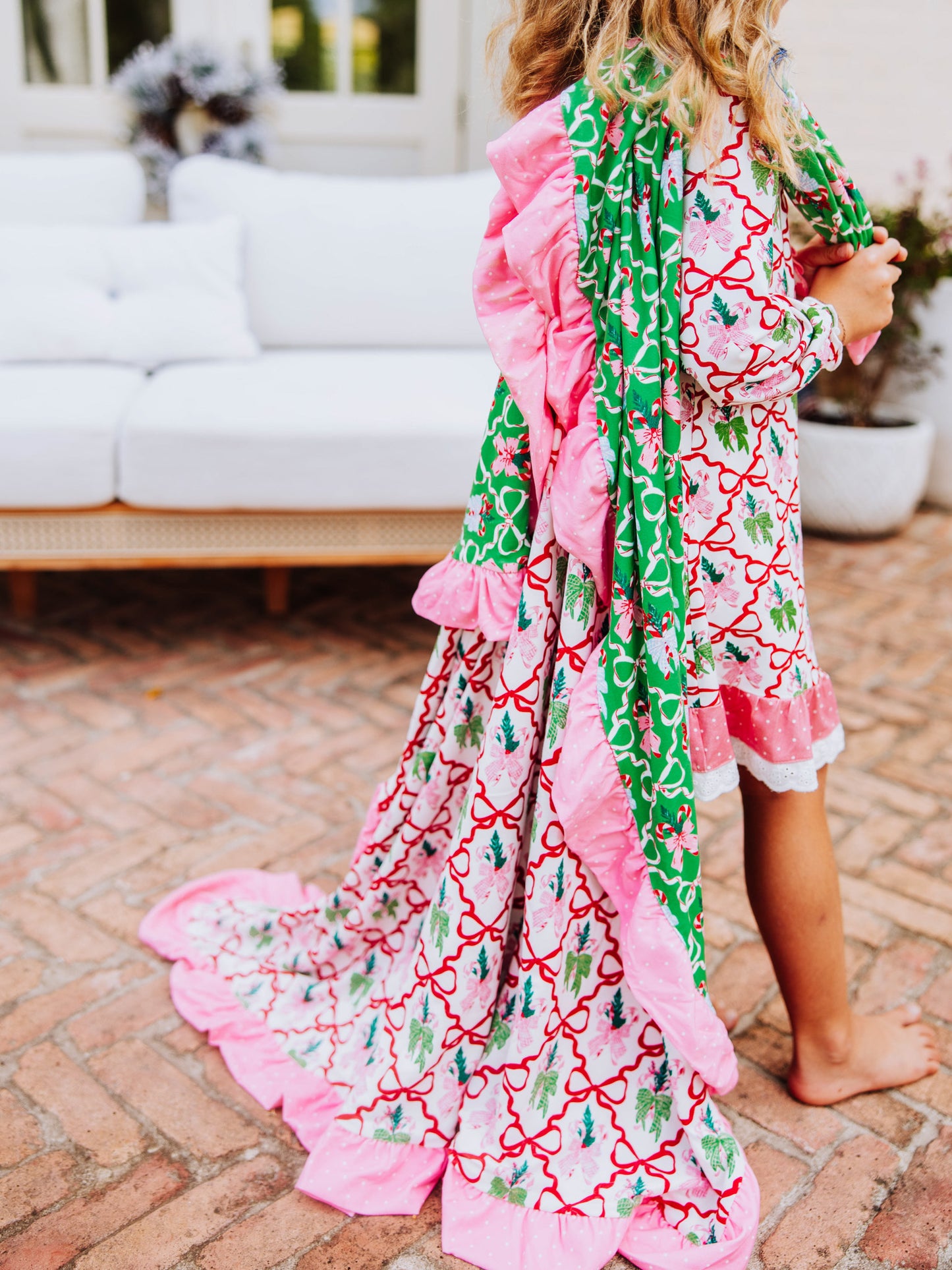 Girl draping a colorful Christmas blanket over shoulder and is wearing a play dress that matches print on blanket. Print is colorful and has red bows throughout pattern.