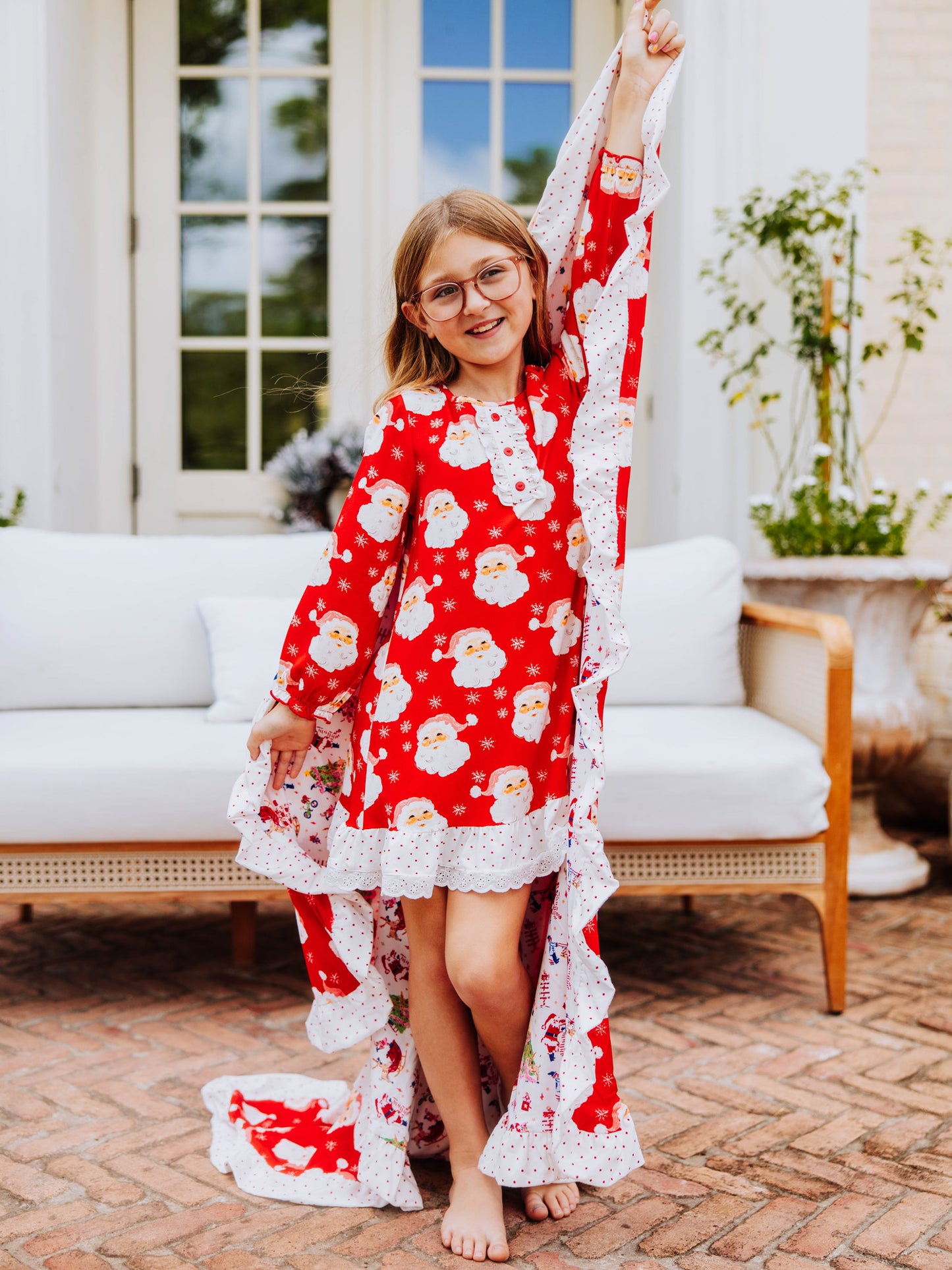 Girl standing outside on brick wearing a red santa dress and holding a matching blanket by the corner in one hand. 