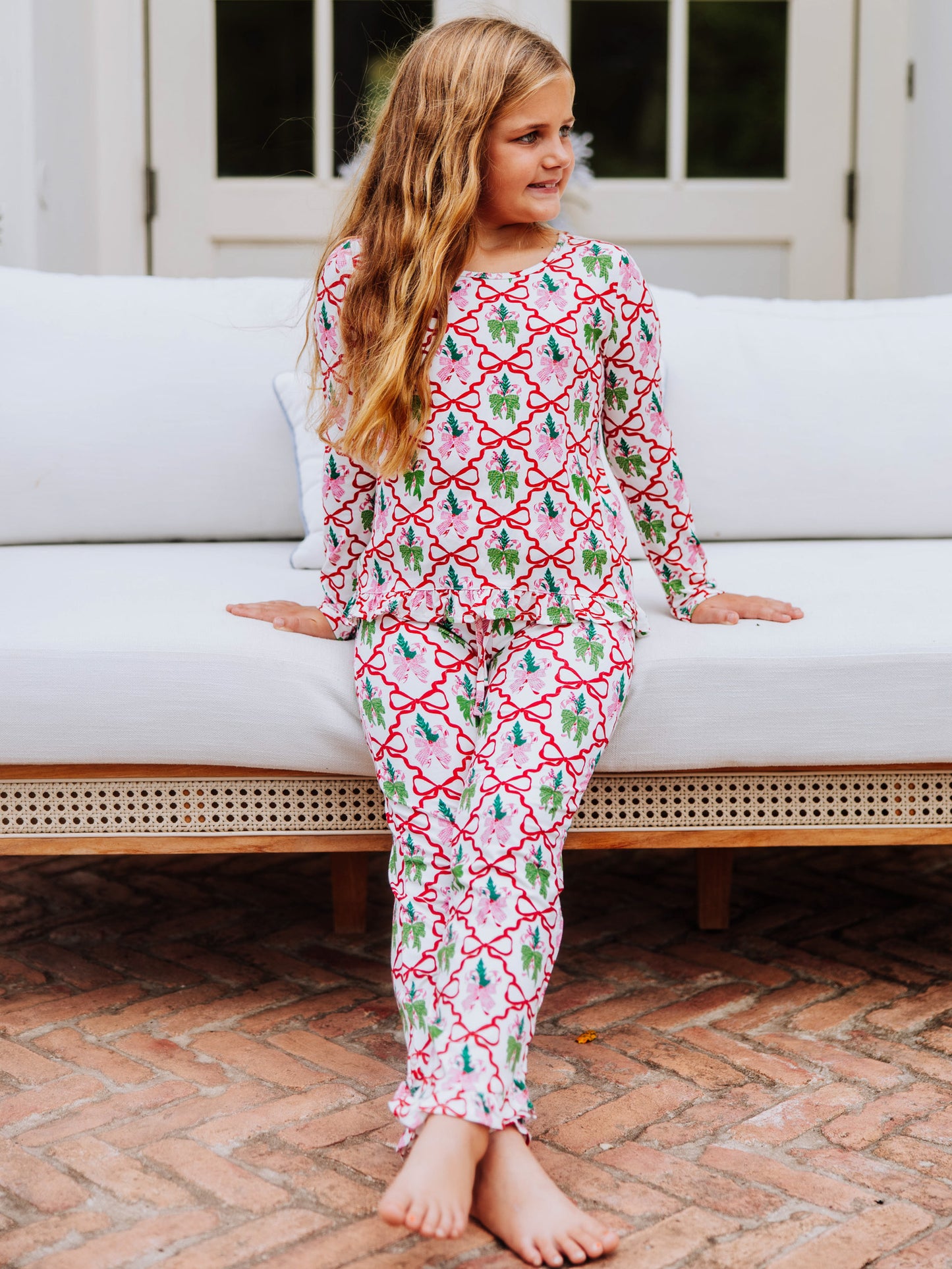 Girl sitting on couch wearing a matching two piece set. The top and bottom have red bows throughout along with some pink and green bow designs. 