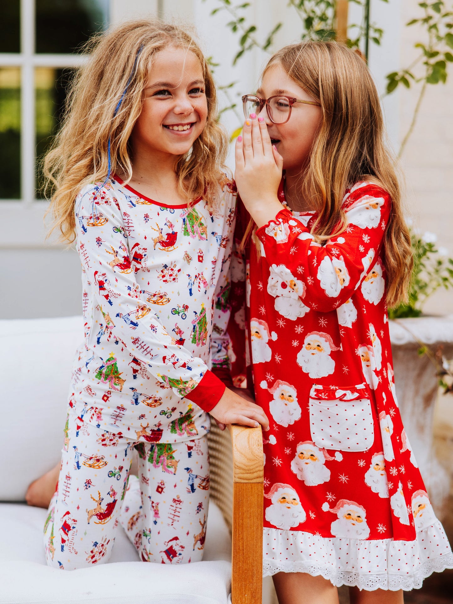 Two girls outside. One on her knees listening while the other is telling her a secret. Left girl is wearing a fitting pair of north pole printed pajamas and the girl on the right is wearing a red santa dres.