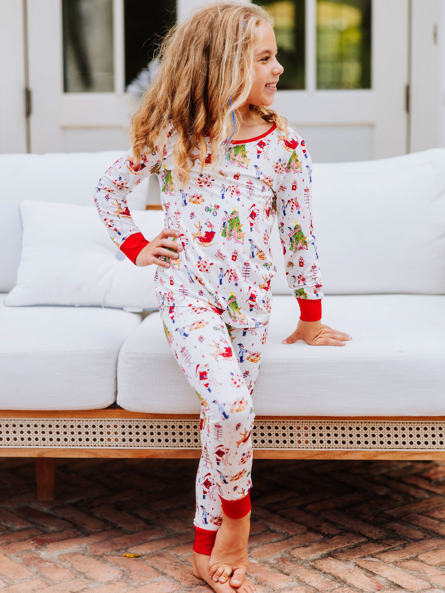 Girl standing with feet crossed outside in front of white couch. Model is wearing a fitted pair of pajamas with a north pole print. 