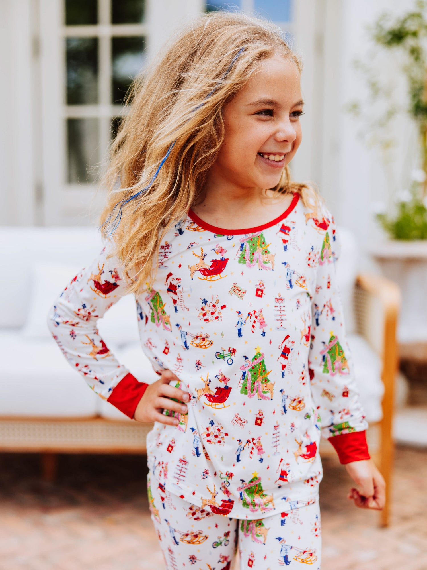 Girl standing with feet crossed outside in front of white couch. Model is wearing a fitted pair of pajamas with a north pole print. 