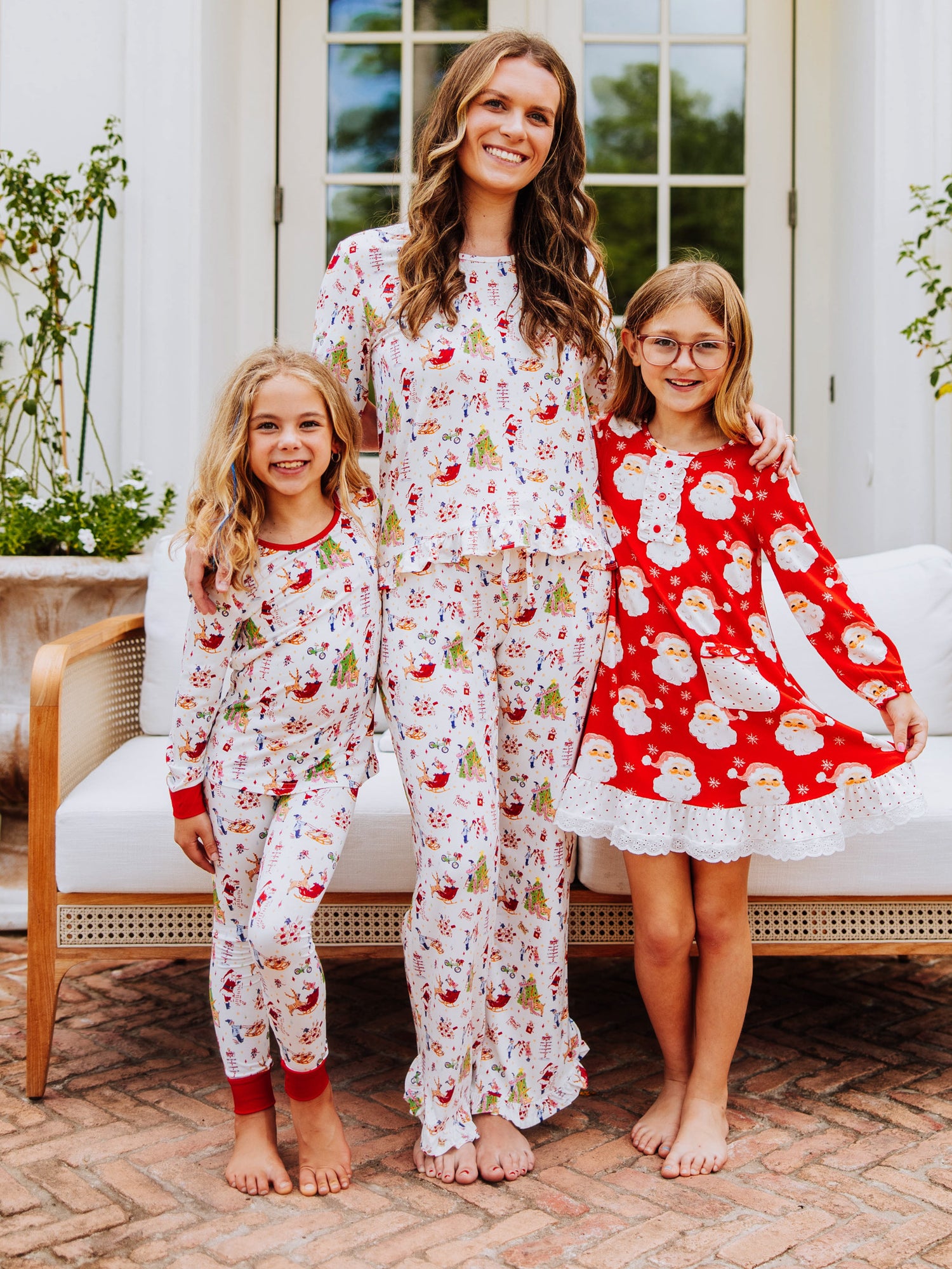 Two girls and a women all standing outside in front of a white couch. Girl and left and women in middle are both wearing matching north pole printed pajamas. Girl on right is wearing a play dress that has Santa heads all over it. 