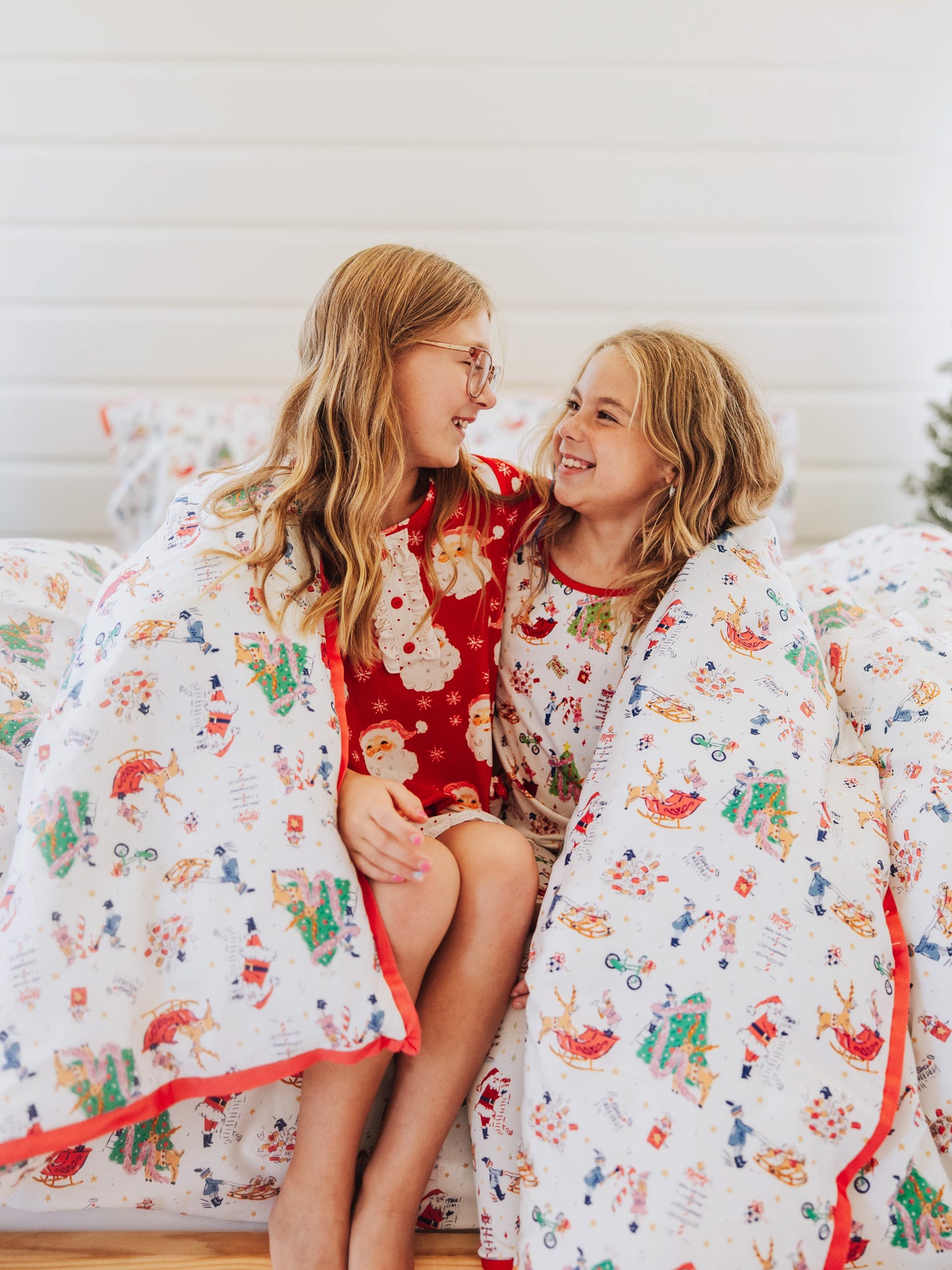 Two girls looking at one another while wrapped up in holiday themed duvet. 