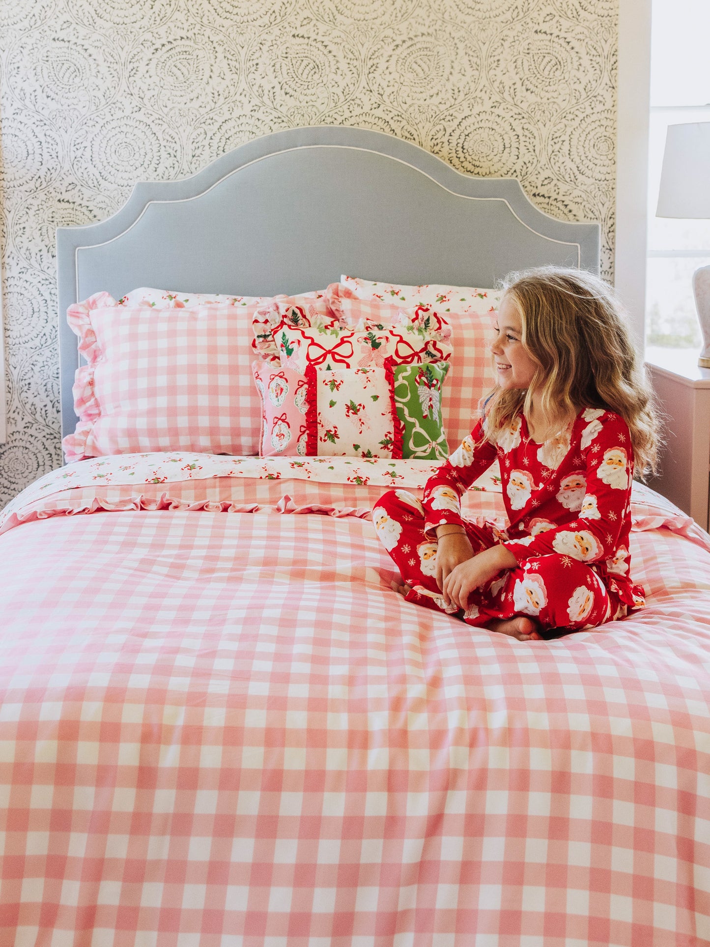 Girl sitting on pink check duvet made bed with holiday themed pillows as well as pink check pillows. 