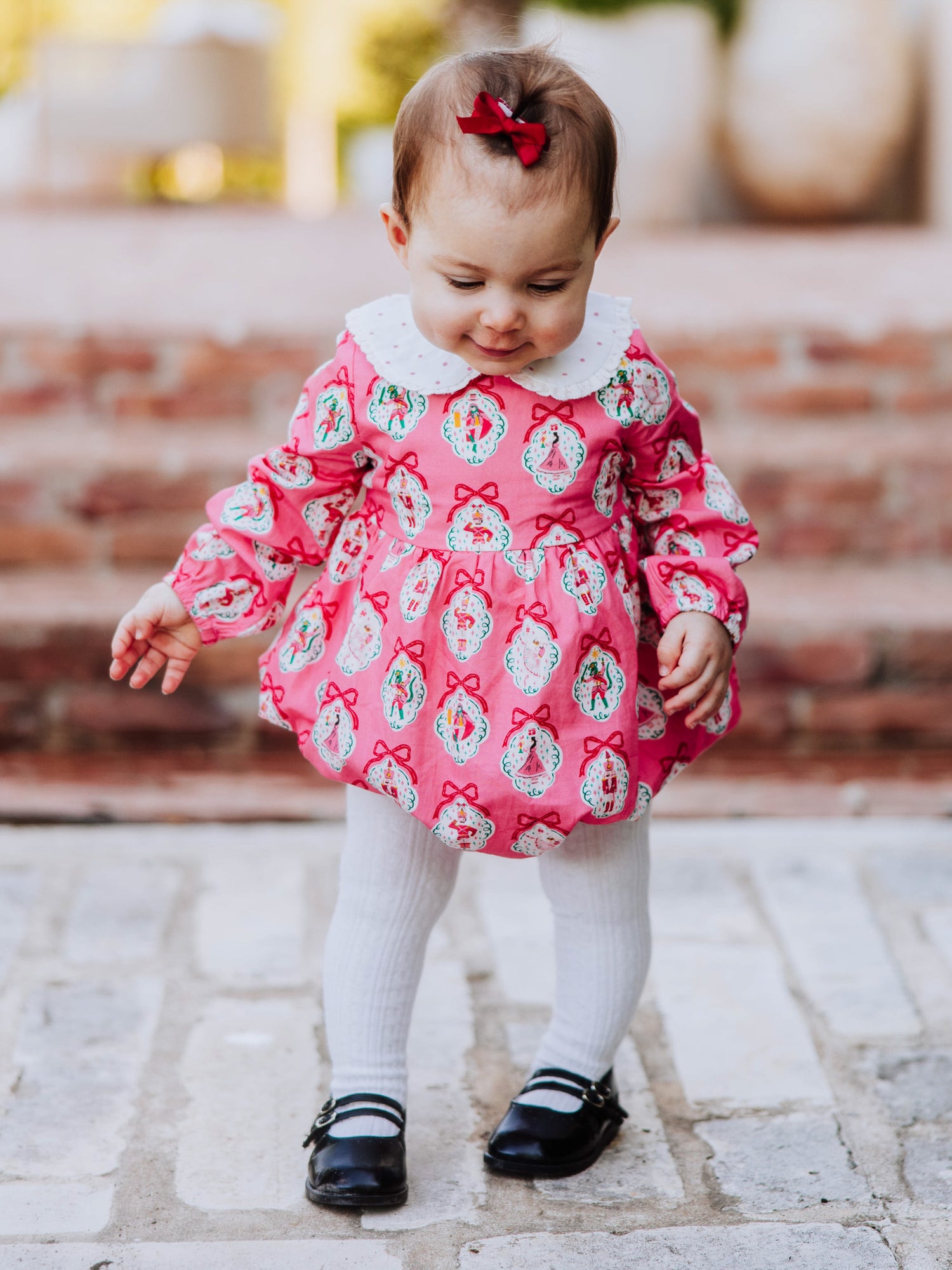 Baby girl outside wearing apink nutcracker print classic bubble with white tights and black mary janes. 