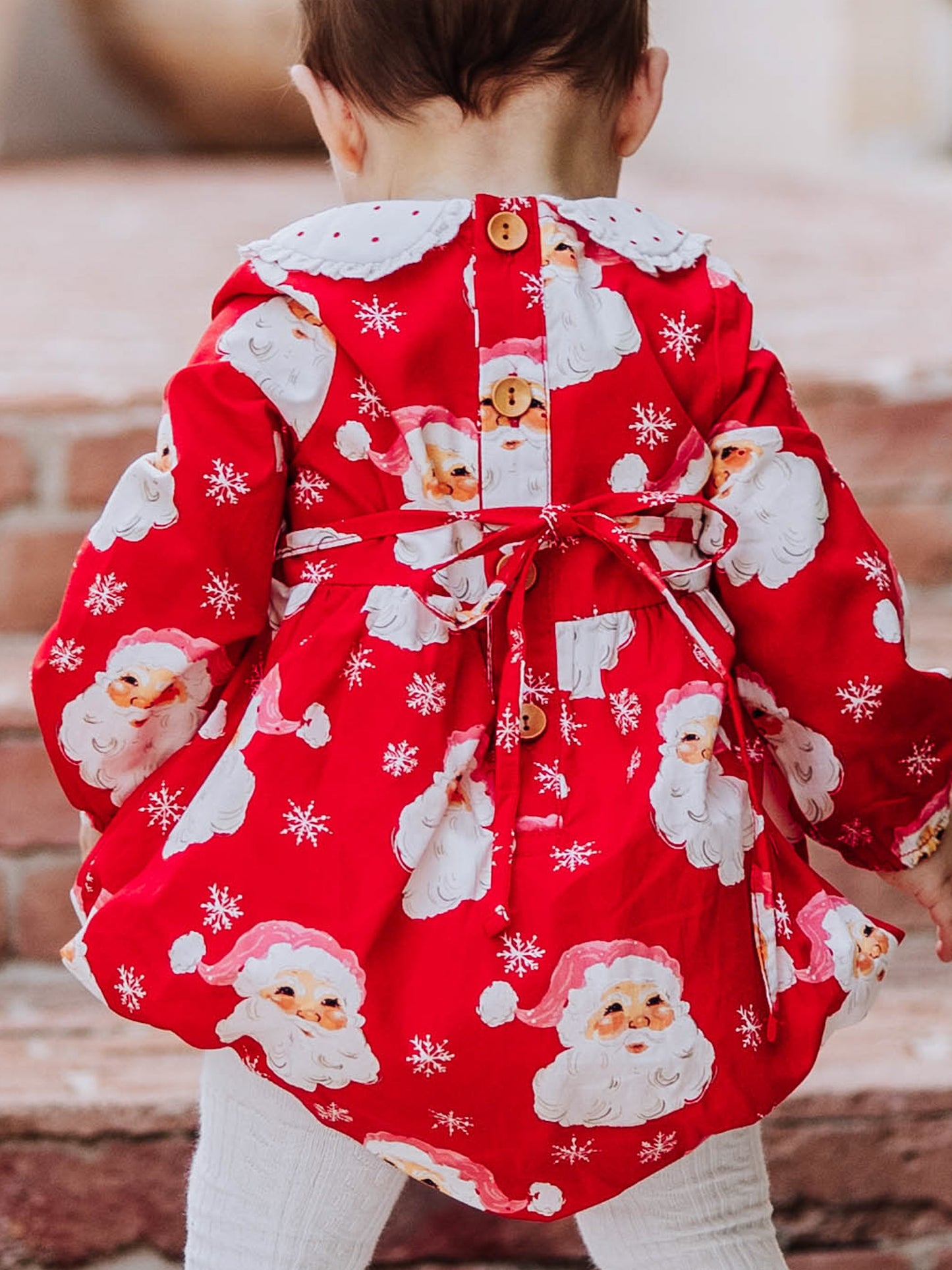 Baby girl outside wearing a jolly santa print classic bubble with white tights and black mary janes. Pic is a close up of back of bubble. You can see the buttons and tie at back. 