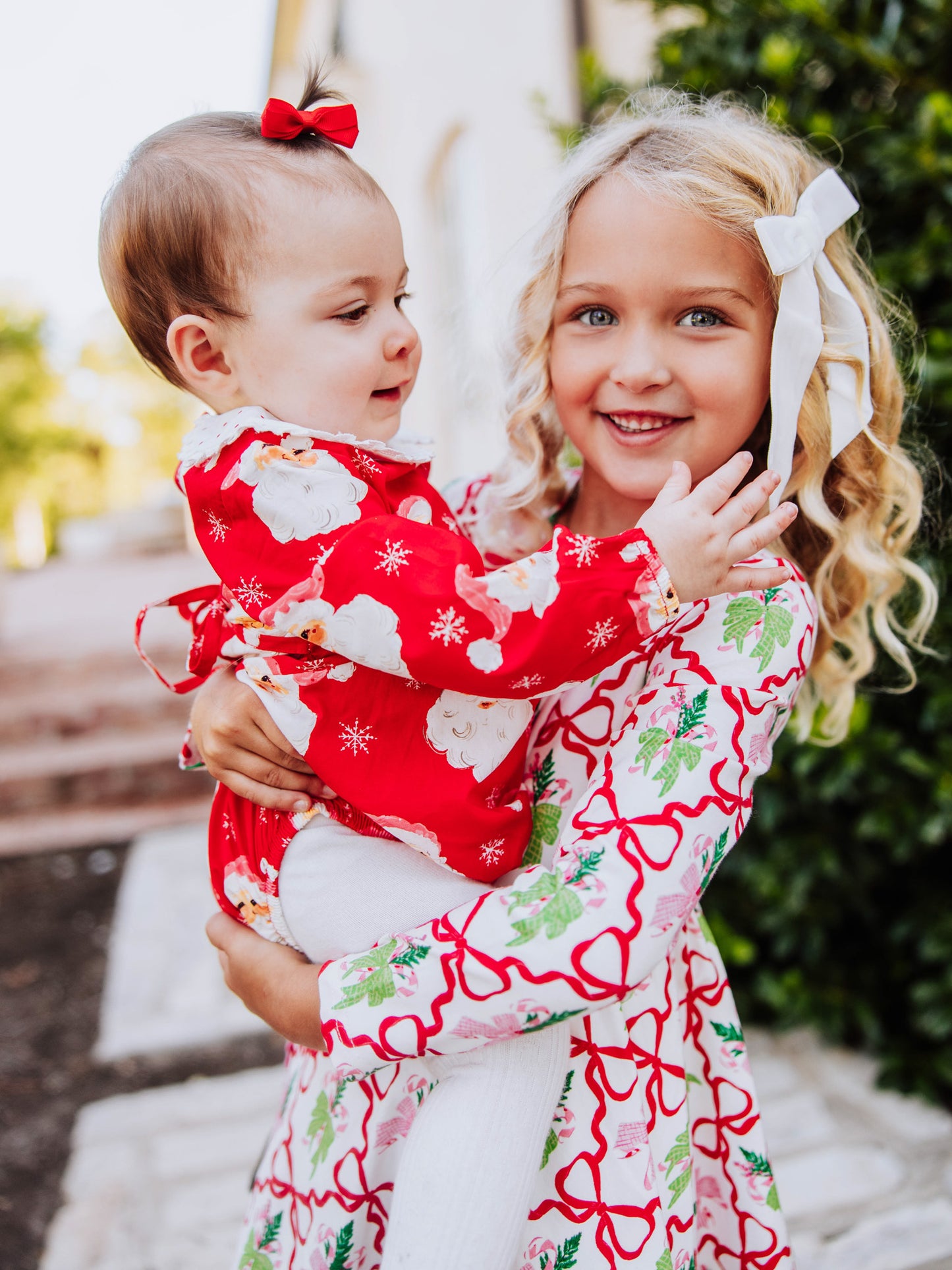 baby girl wearing a red jolly santa bubble and is being held by a girl who is wearing a holiday themed dress. 