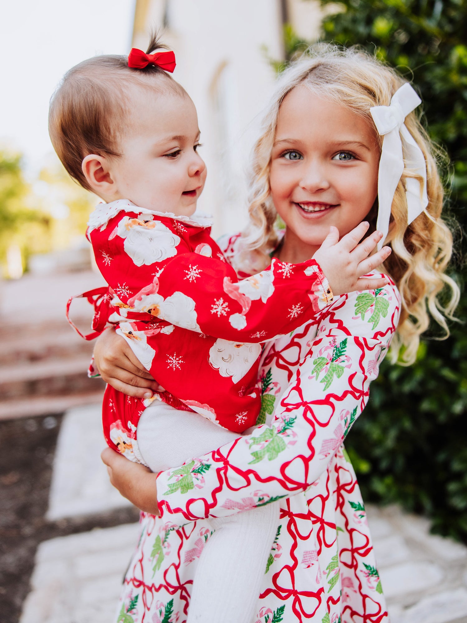 baby girl wearing a red jolly santa bubble and is being held by a girl who is wearing a holiday themed dress. 
