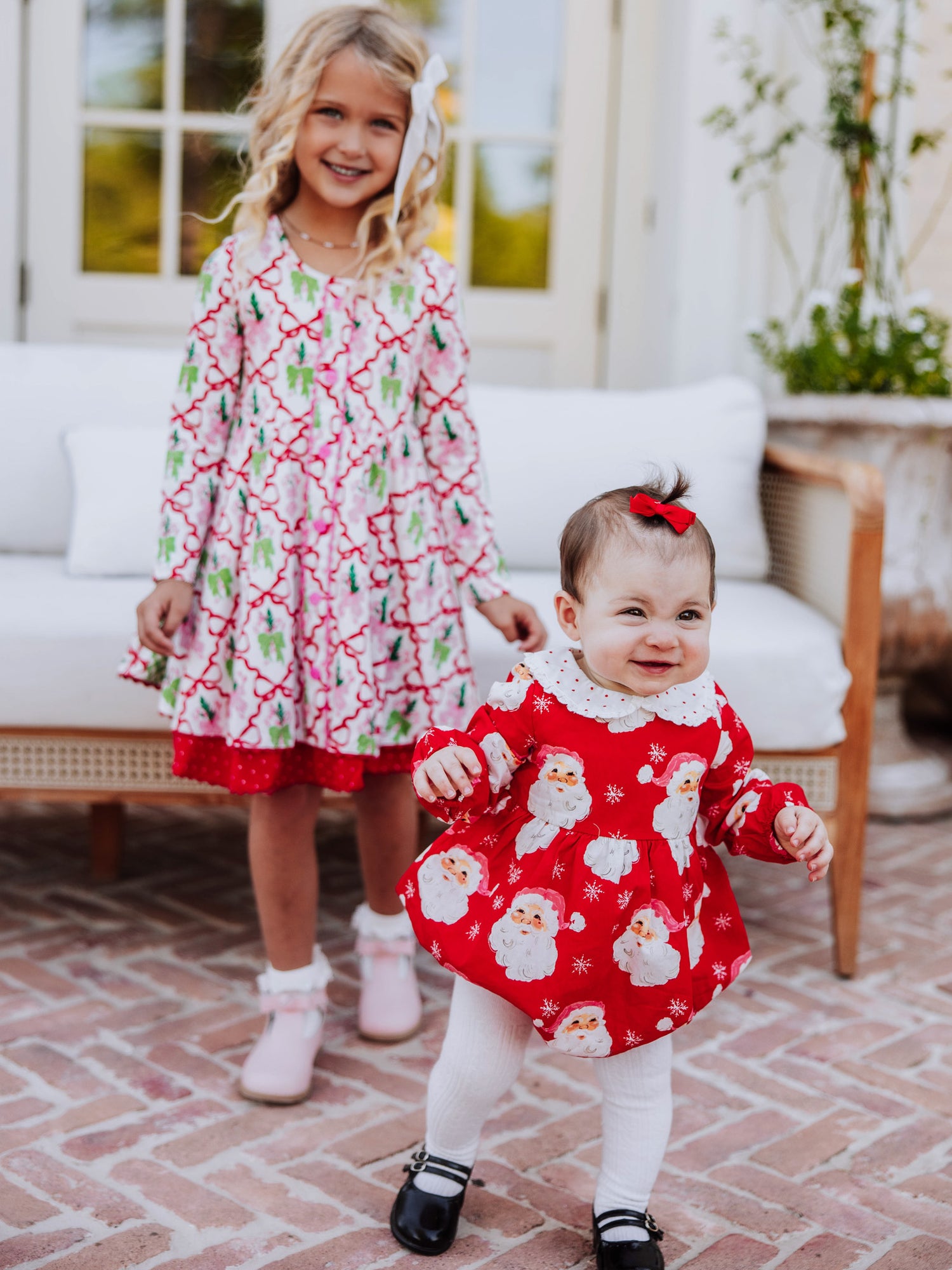 Baby girl outside wearing a jolly santa print classic bubble with white tights and black mary janes. Girl standing behind her in front of a couch and is wearing a holiday themed dress. 