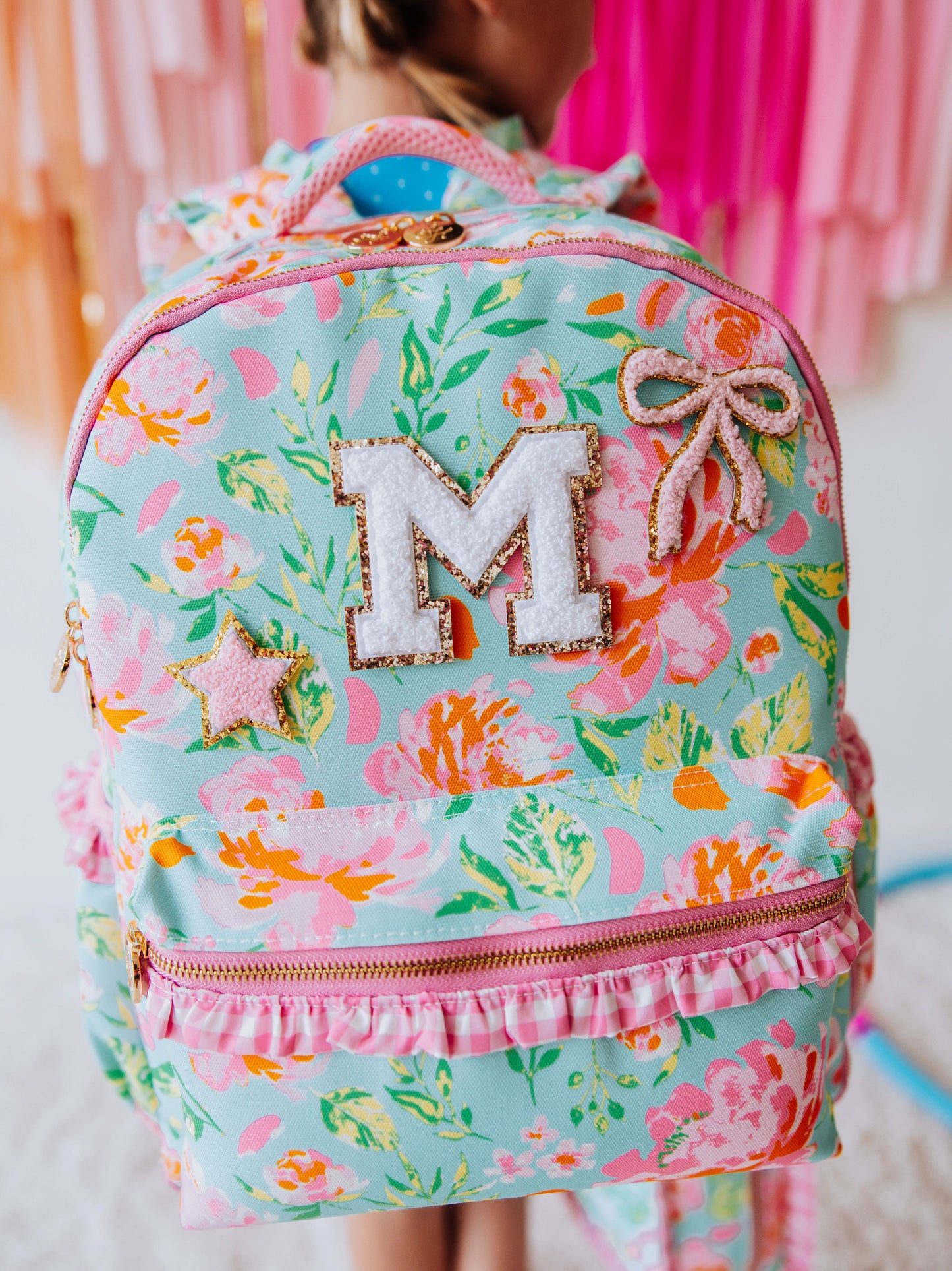 Girl wearing a colorful, floral backpack that has white and pink patches on the front