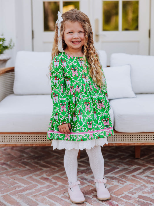 Girl standing in front of a white couch wearing a holiday themed dress with white  tights and pink buckle strap boots. 