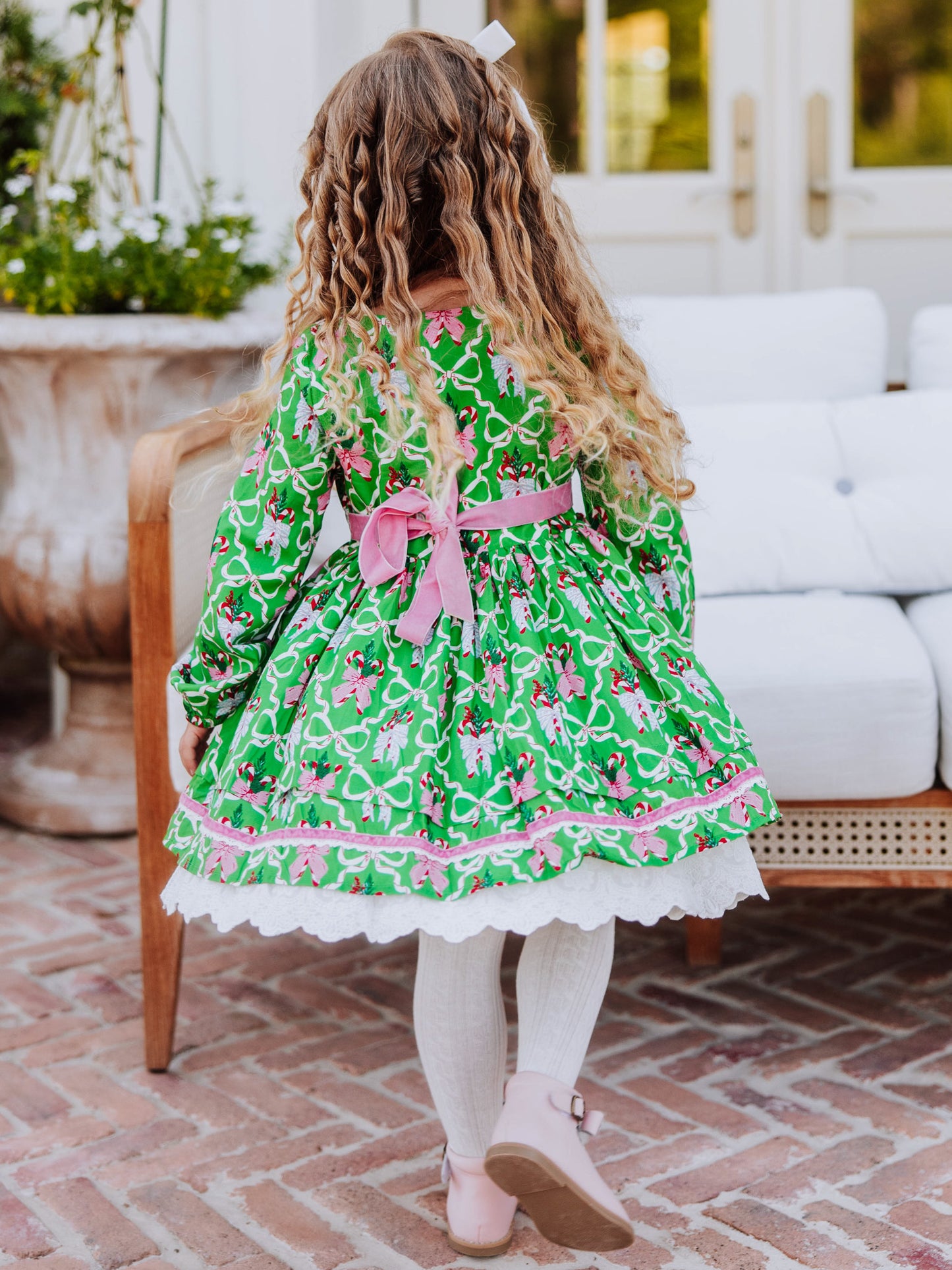 Girl standing in front of a white couch wearing a holiday themed dress with white  tights and pink buckle strap boots. 