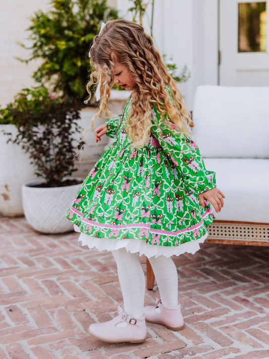 Girl standing in front of a white couch wearing a holiday themed dress with white  tights and pink buckle strap boots. 