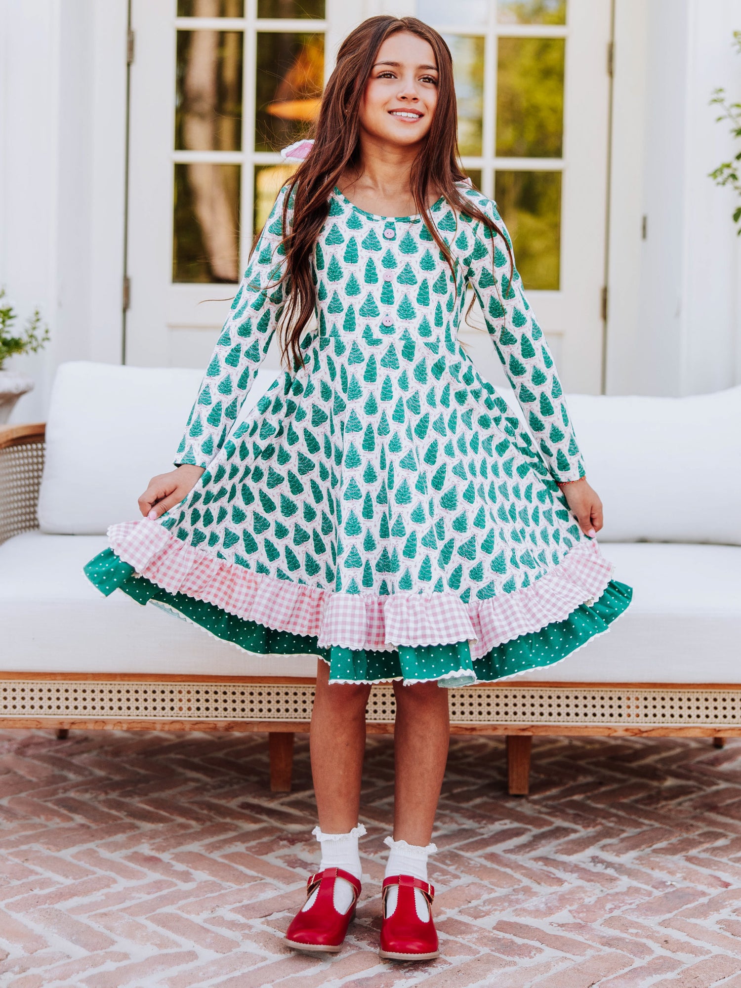 Girl standing outside in front of couch holding both sides of her Christmas tree dress that has double ruffles. 
