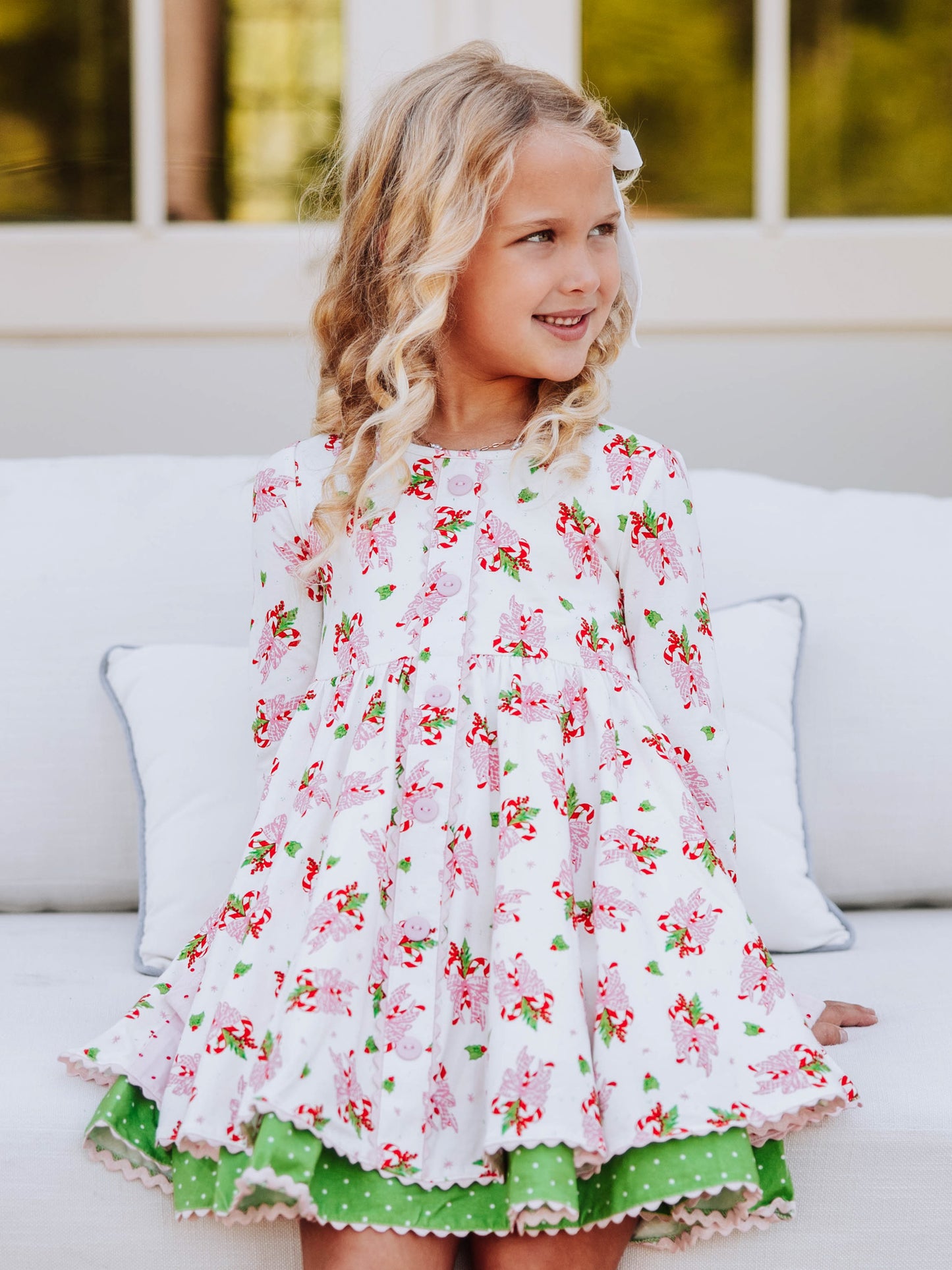 Girl is sitting on white couch wearing a holiday themed dress. 