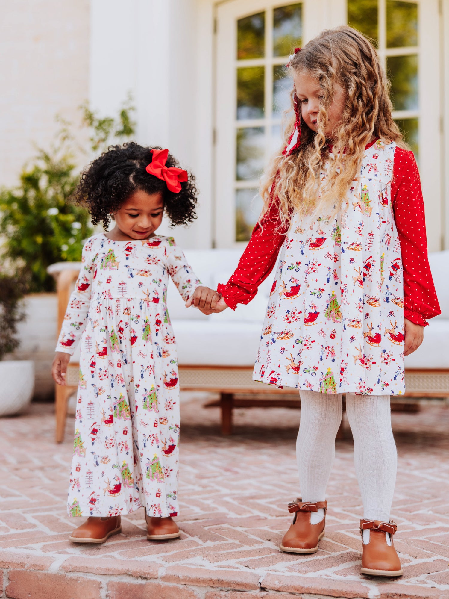 Two girls holding hands and are wearing matching printed holiday clothing. One is in a retro jumper and the other is in a leggy romper. 