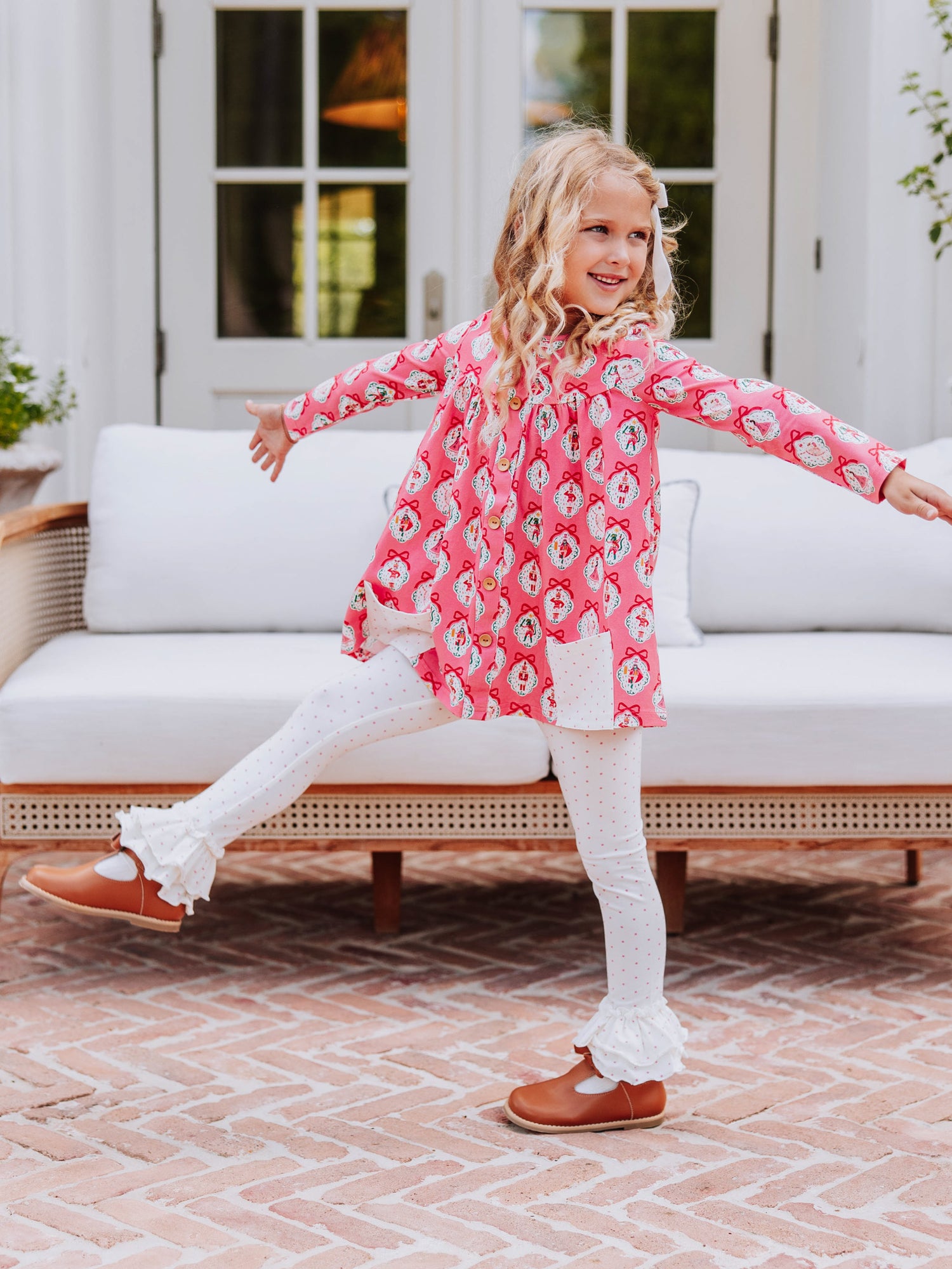 Girl kicking leg out and holding arms out wearing a pink holiday themed top with white leggings with pink polka dots. 
