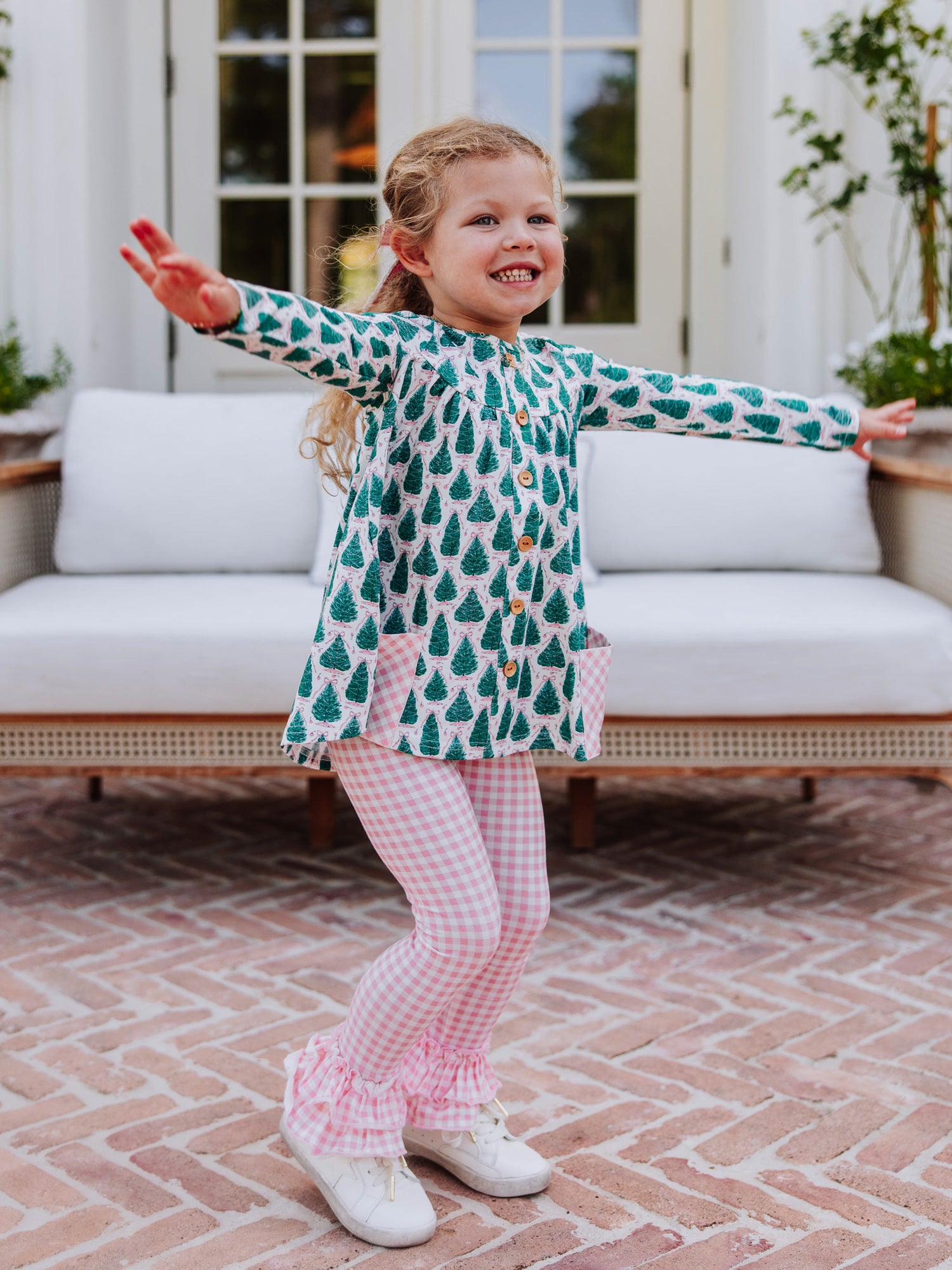 Girl holding arms out wearing a Christmas tree themed out to play set with pink gingham bottoms and white tennis shoes. 