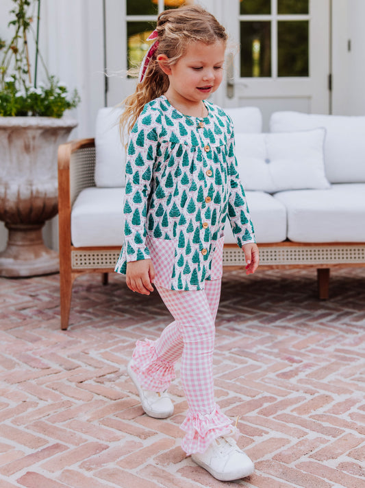 Girl holding arms out wearing a Christmas tree themed out to play set with pink gingham bottoms and white tennis shoes. 