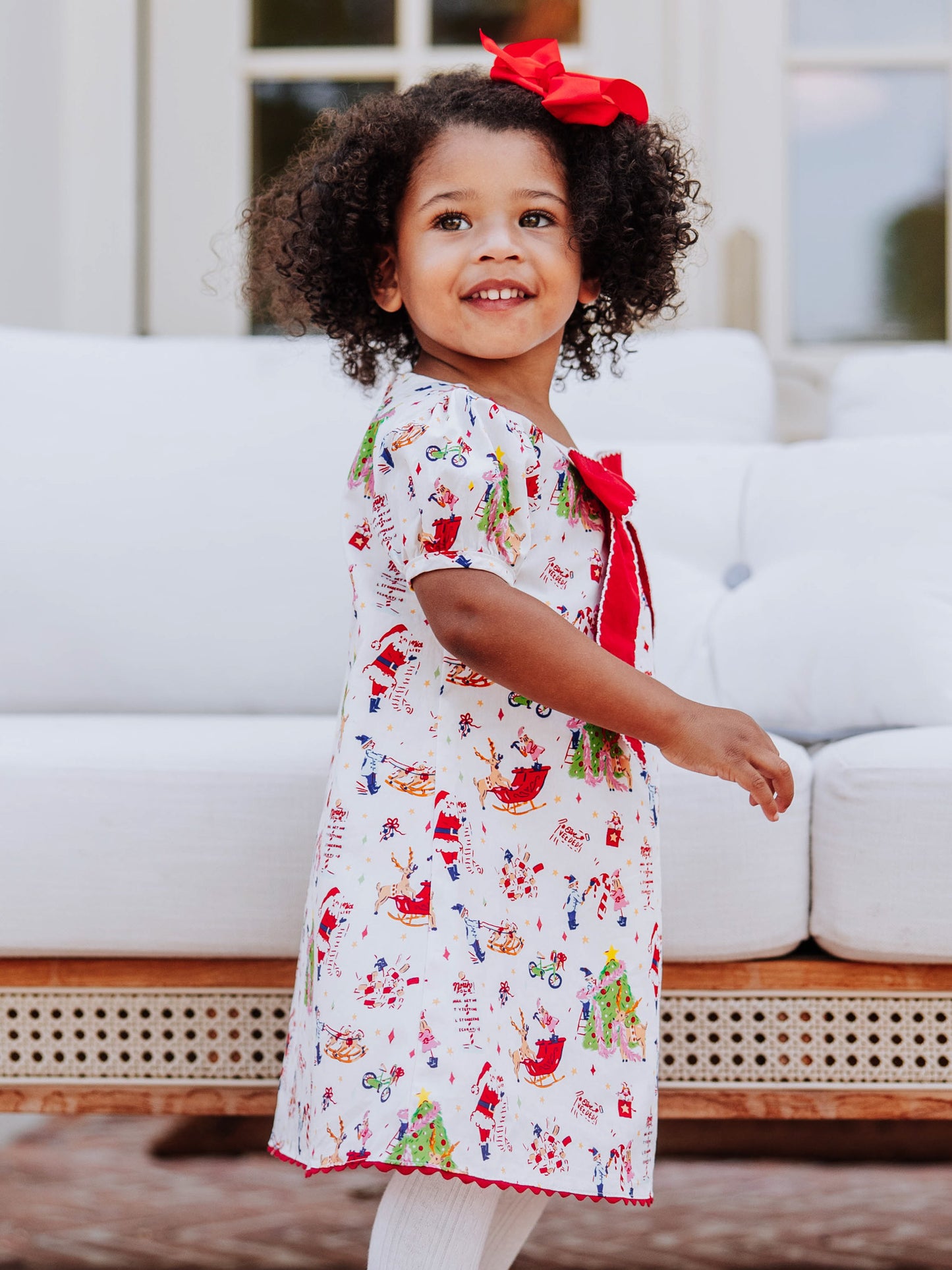 Model posing in front of a white couch wearing a holiday printed dress with white tights and brown buckle boots. 