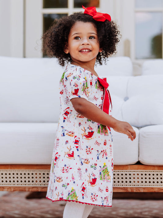 Model posing in front of a white couch wearing a holiday printed dress with white tights and brown buckle boots. 