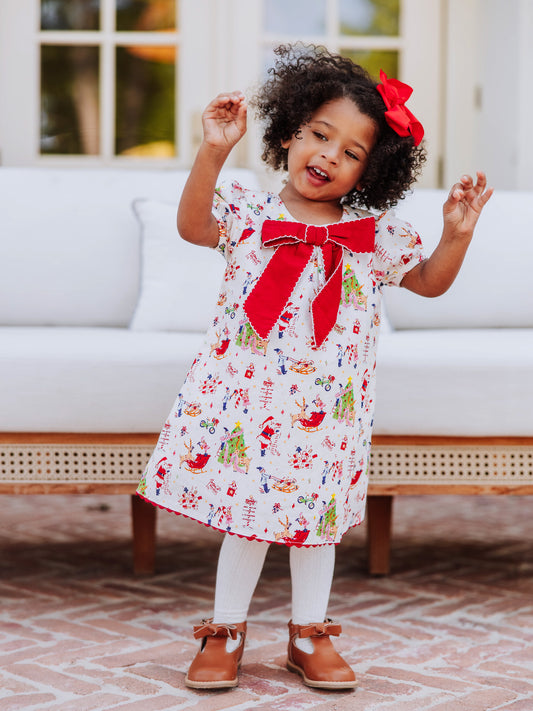 Model posing in front of a white couch wearing a holiday printed dress with white tights and brown buckle boots. 