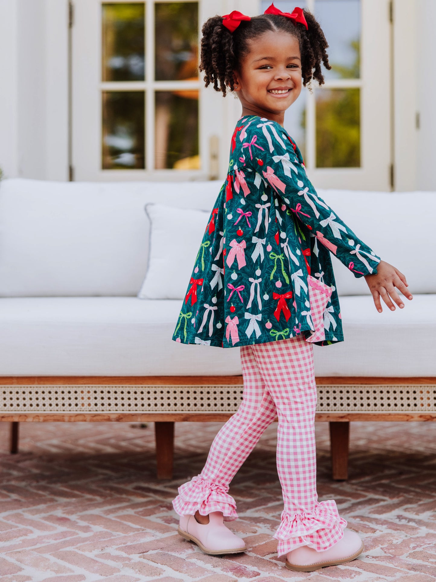 Girl wearing a holiday themed two piece set. Top has colorful bows and bottoms and accent pockets are pink gingham. 