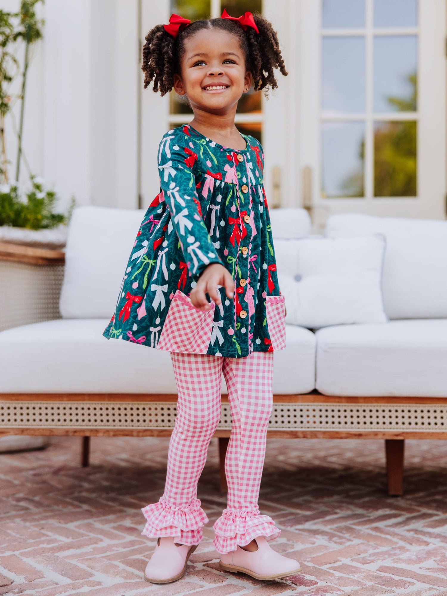 Girl wearing a holiday themed two piece set. Top has colorful bows and bottoms and accent pockets are pink gingham. 