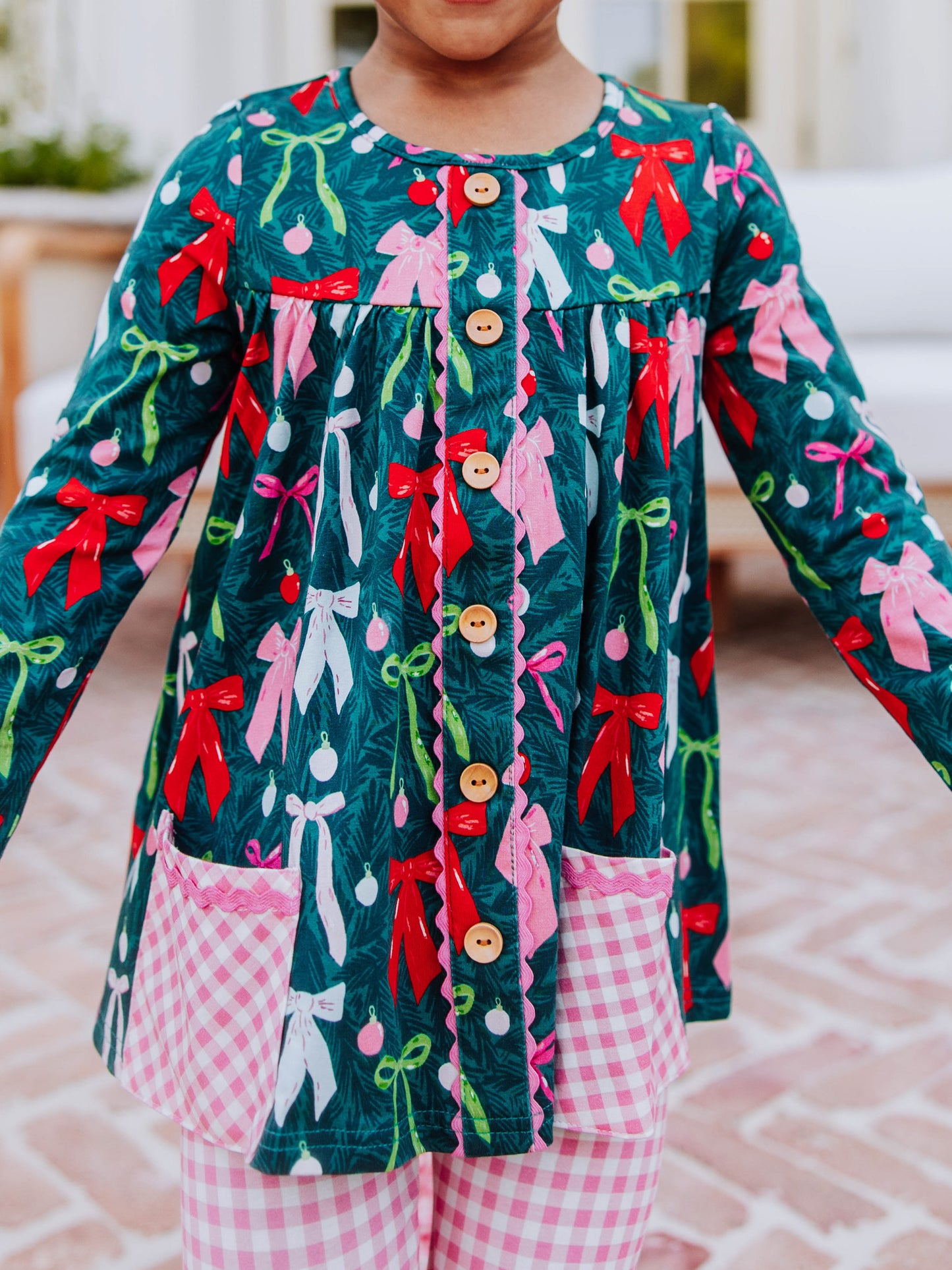 Girl wearing a holiday themed two piece set. Top has colorful bows and bottoms and accent pockets are pink gingham. 