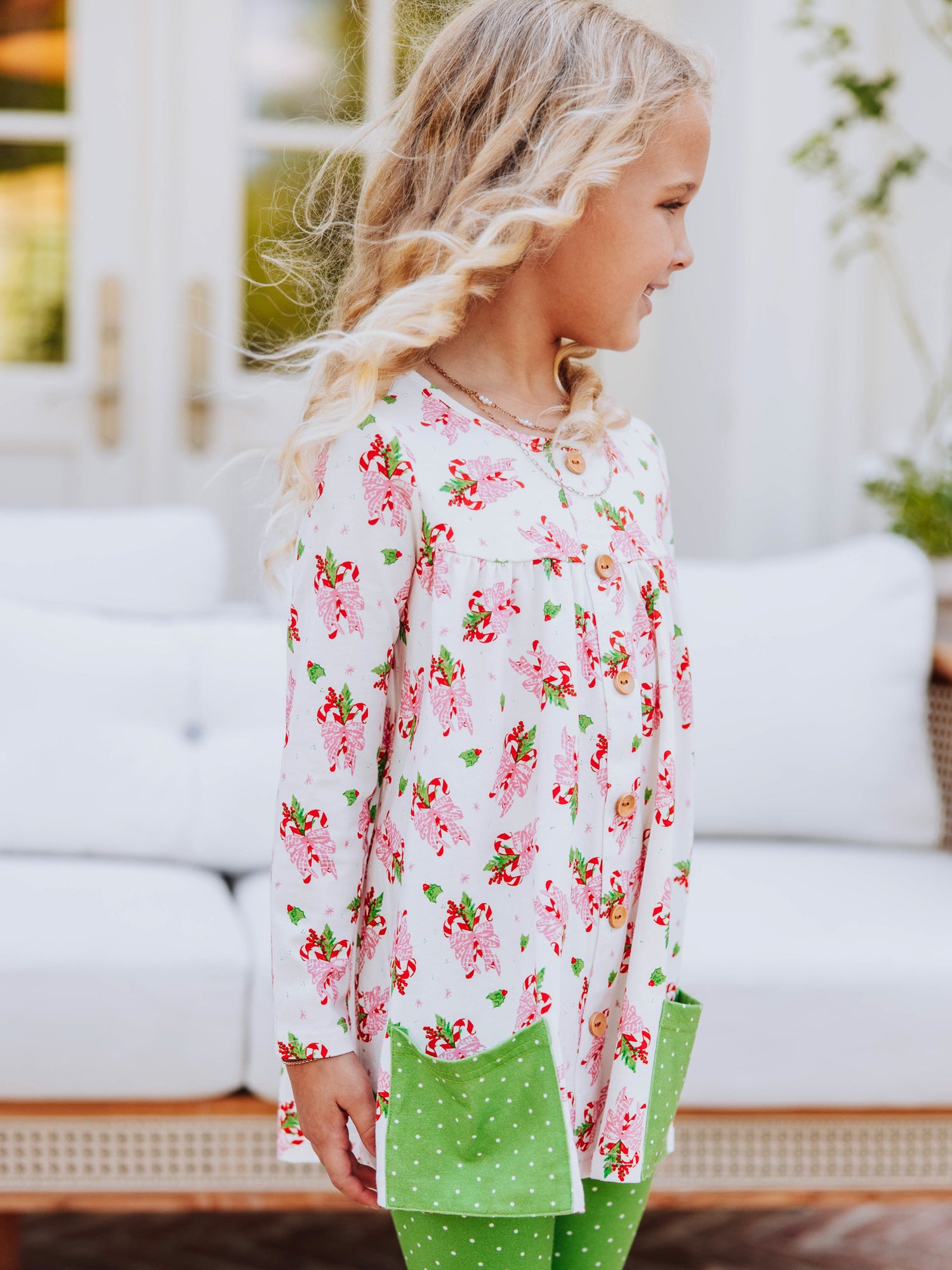 Girl standing outside in front of couch wearing a holiday themed top with green leggings with white polka dots that have ruffles at bottom. 