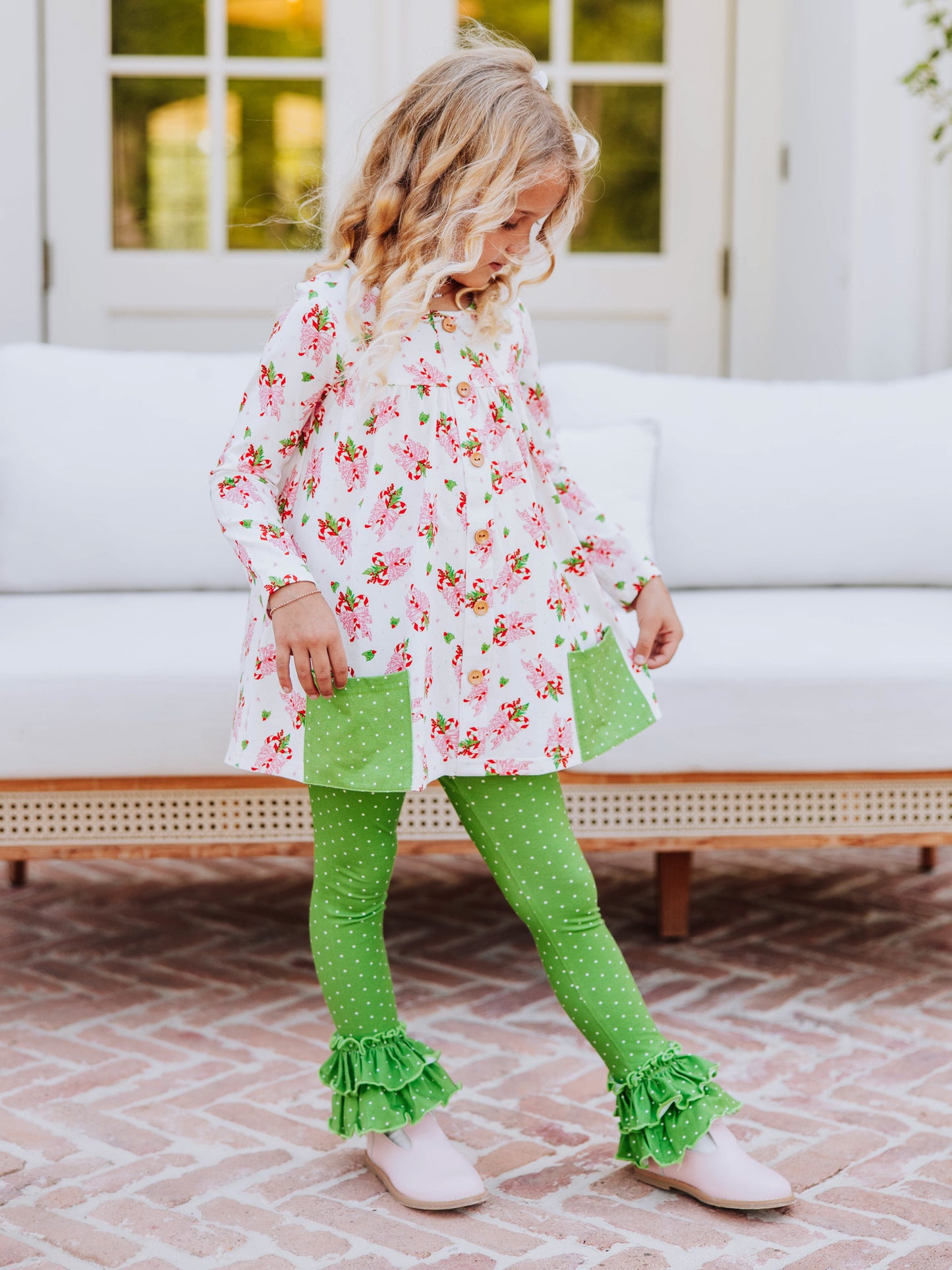 Girl standing outside in front of couch wearing a holiday themed top with green leggings with white polka dots that have ruffles at bottom. 
