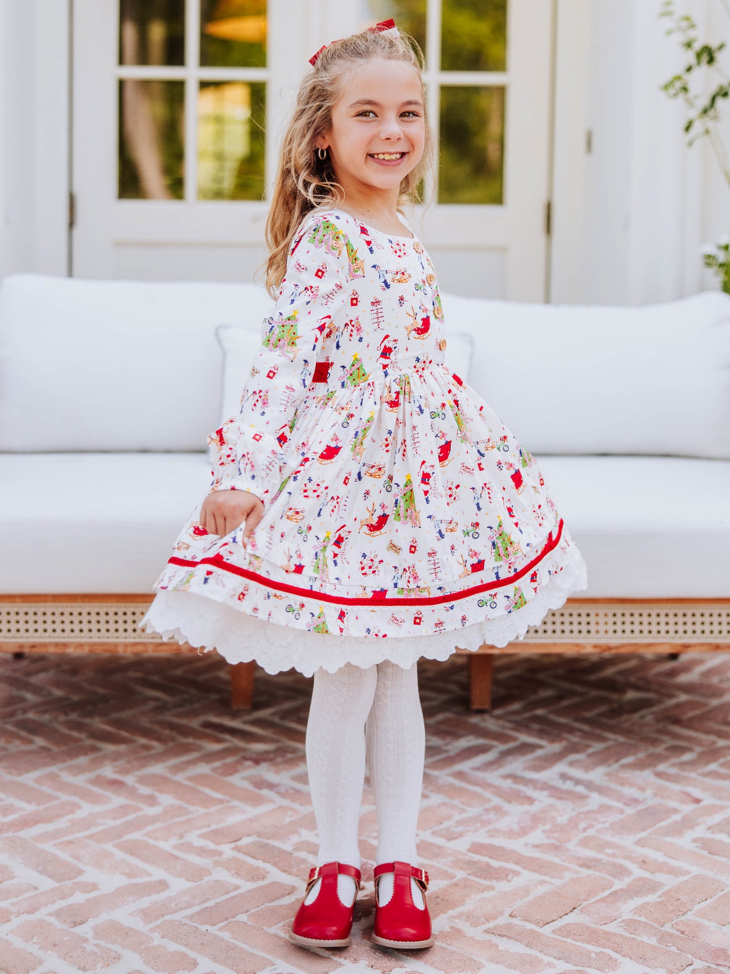 Girl wearing a holiday themed dress and is holding it on both corners to see. Girl also wearing white tights with red mary janes. 
