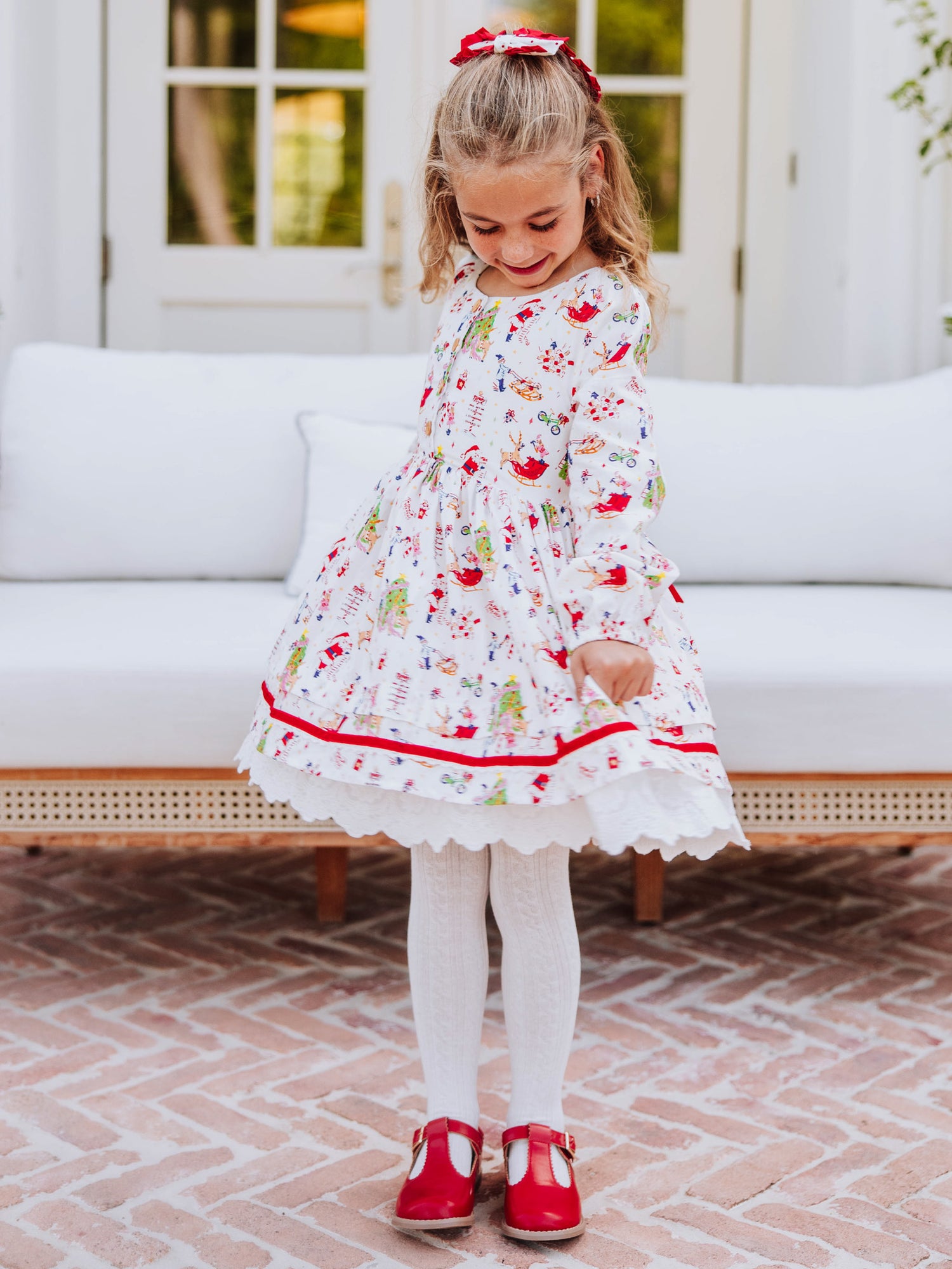 Girl wearing a holiday themed dress and is holding it on both corners to see. Girl also wearing white tights with red mary janes. 