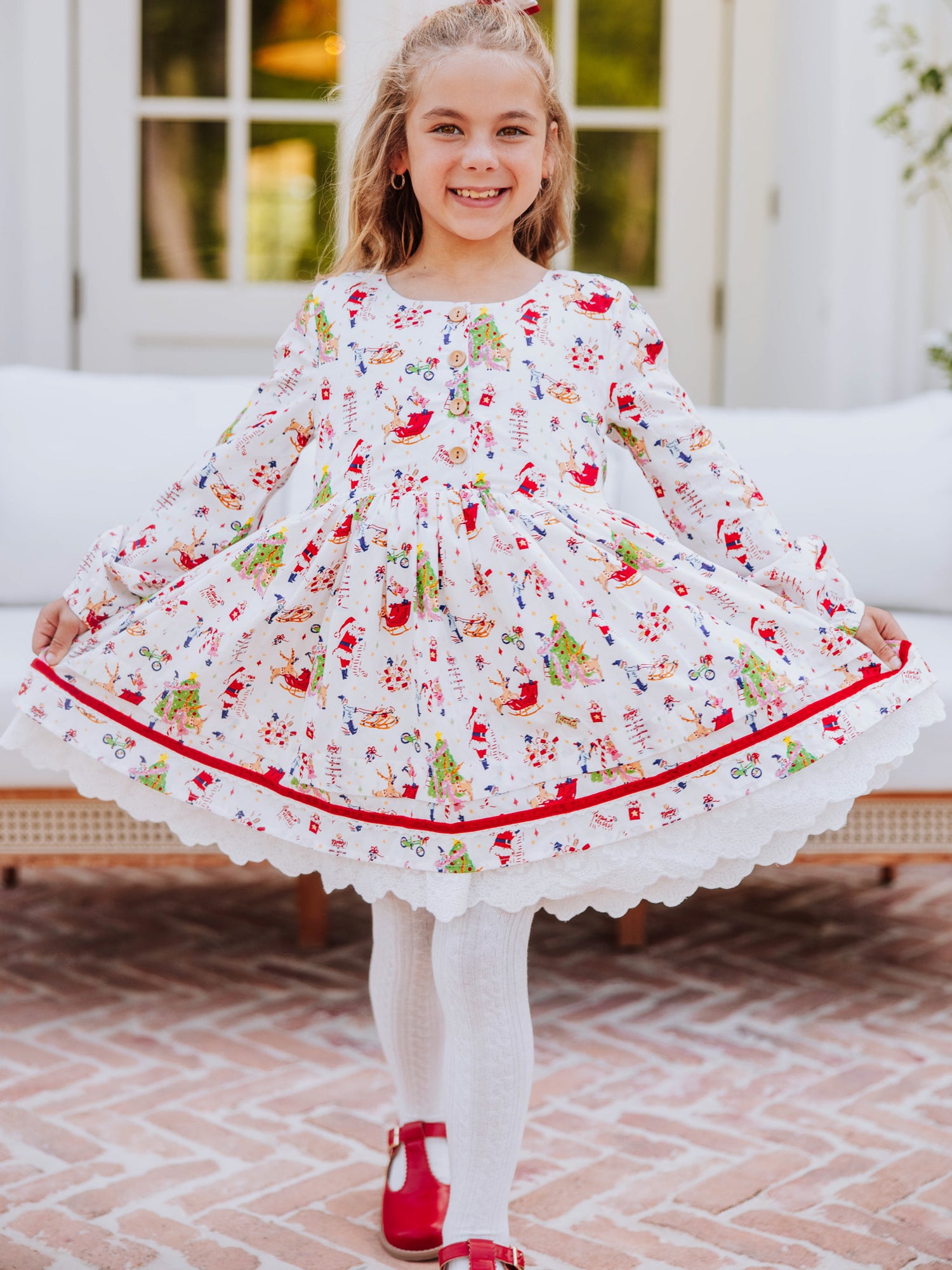 Girl wearing a holiday themed dress and is holding it on both corners to see. Girl also wearing white tights with red mary janes. 